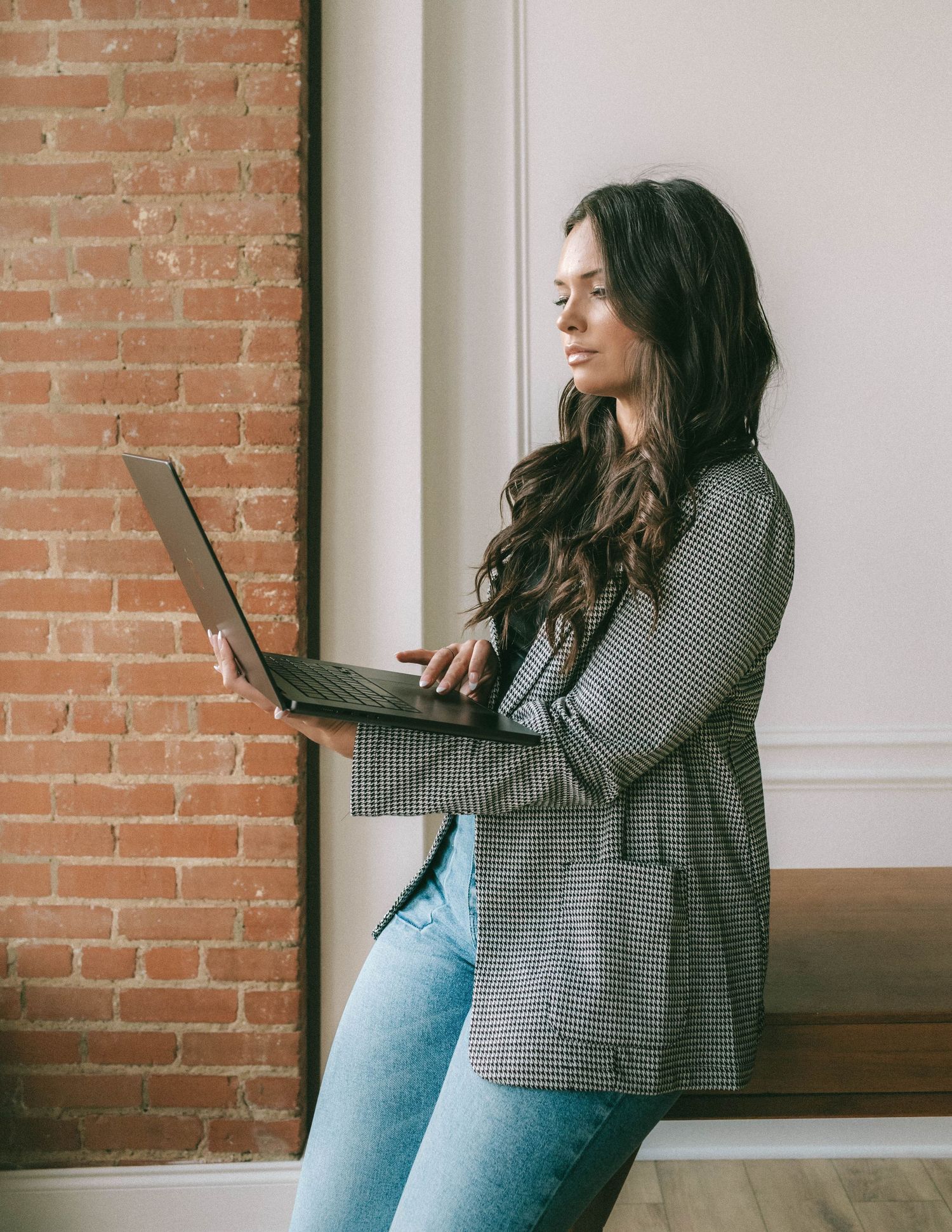 Woman in striped top and jeans leans casually against brick wall while holding laptop computer.