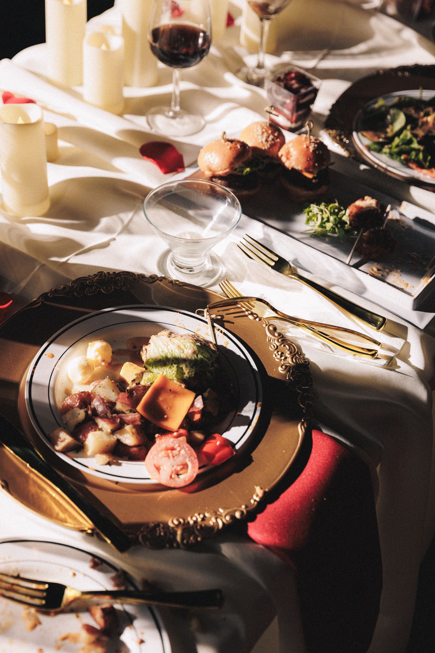 Elegant wedding reception dinner table with gold chargers, candles, red wine, and gourmet food.