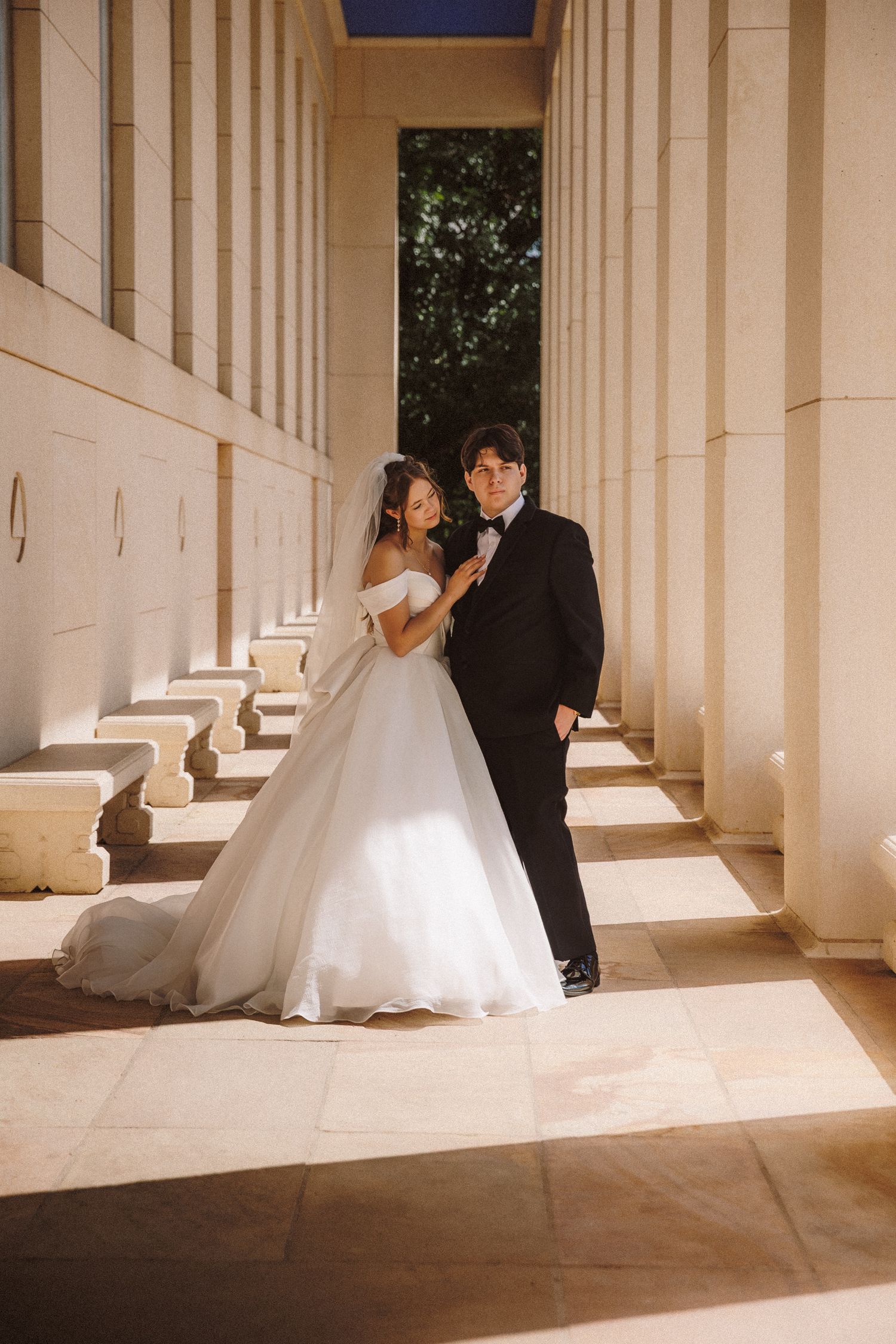 Bride and groom posing between sunlit stone columns in classic wedding portrait outdoors.