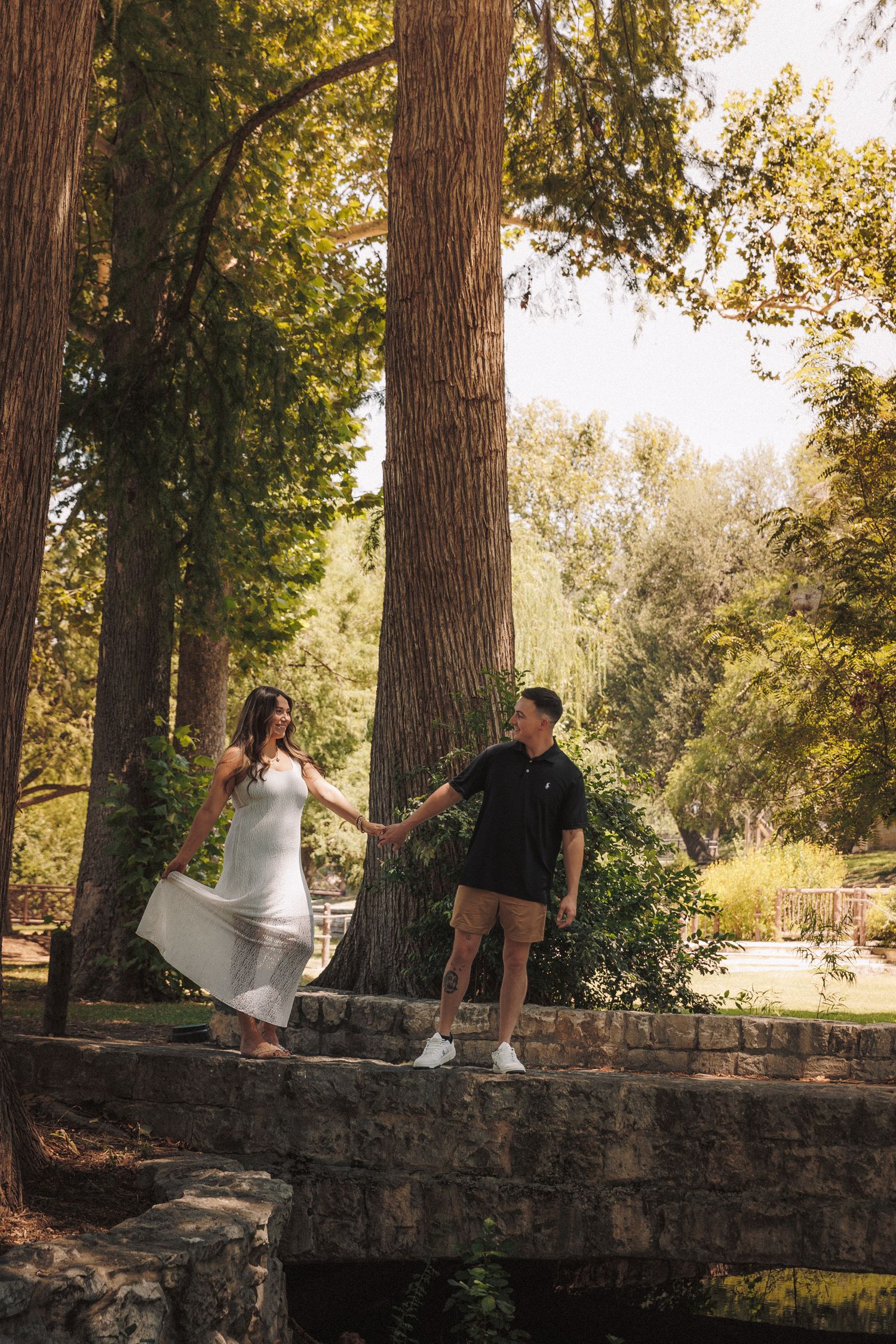 A couple in white and black outfits share a playful moment on a rustic stone wall surrounded by tall trees.