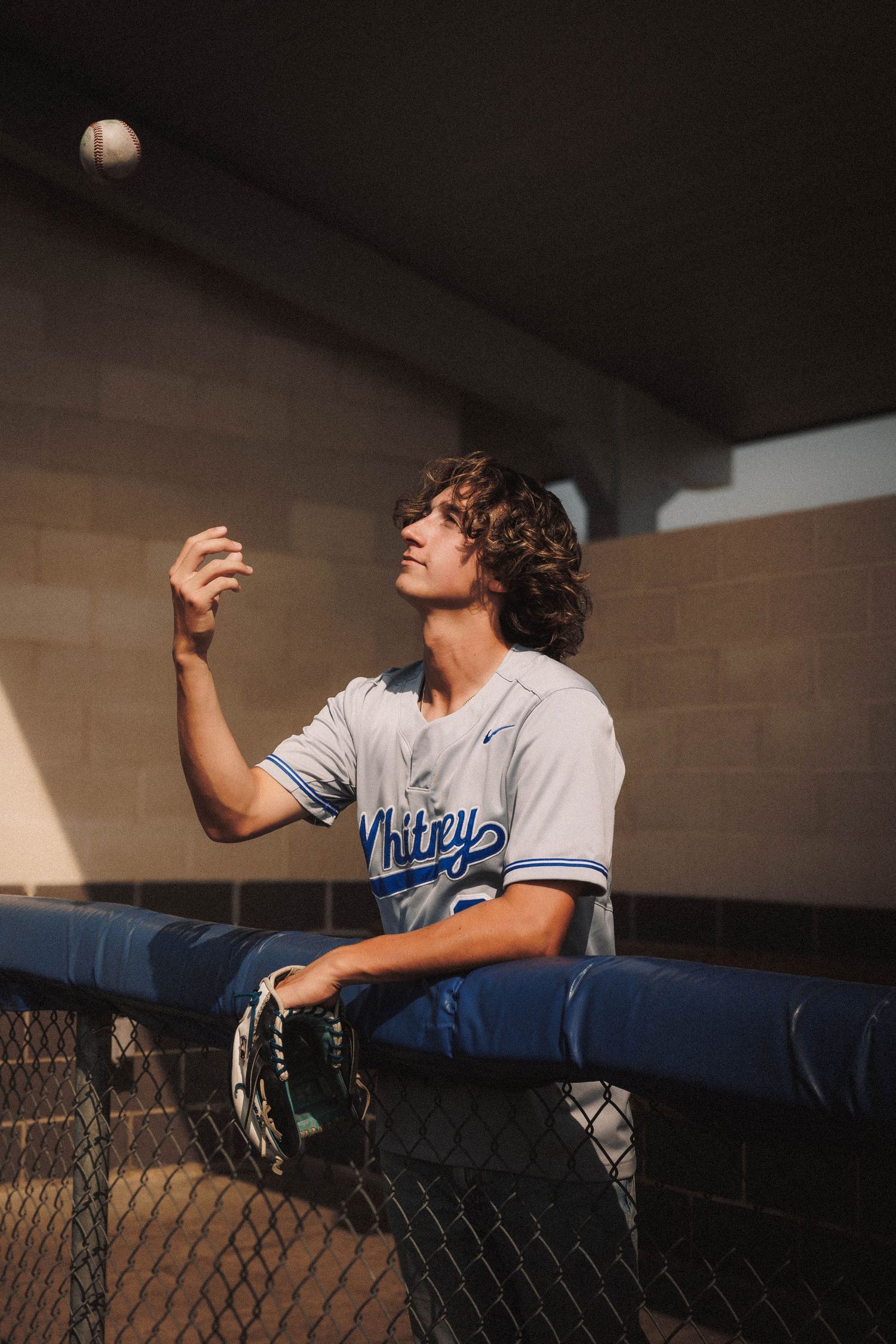 Baseball player in white uniform leans against chain link fence during sunset at baseball field.