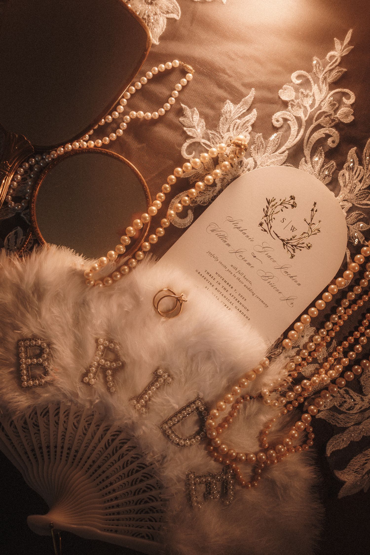 Elegant bridal flat lay with pearl necklaces, wedding invitation, feather fan, and diamond ring on lace fabric.