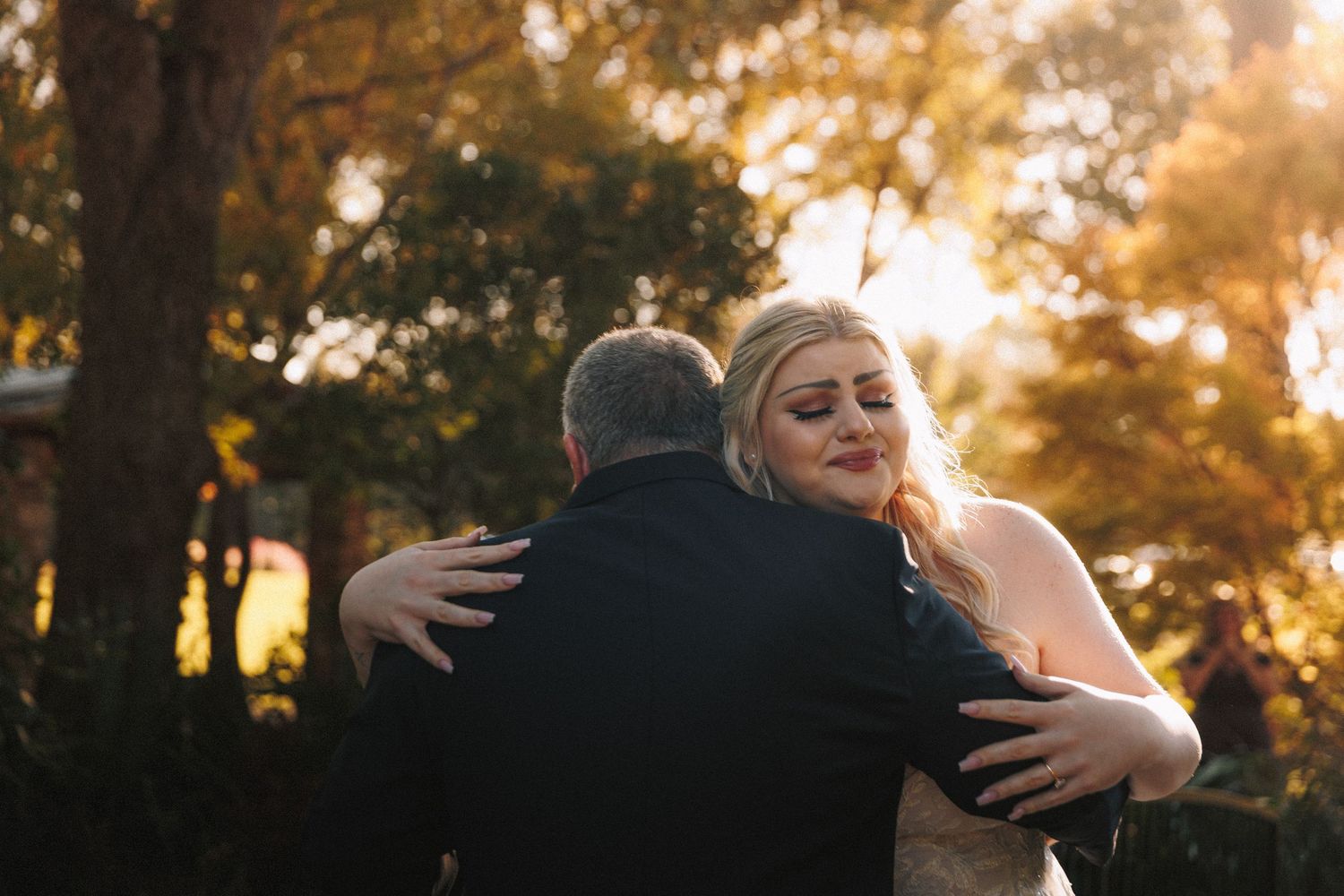 Emotional bride hugging guest in golden sunset light outdoors at wedding reception.