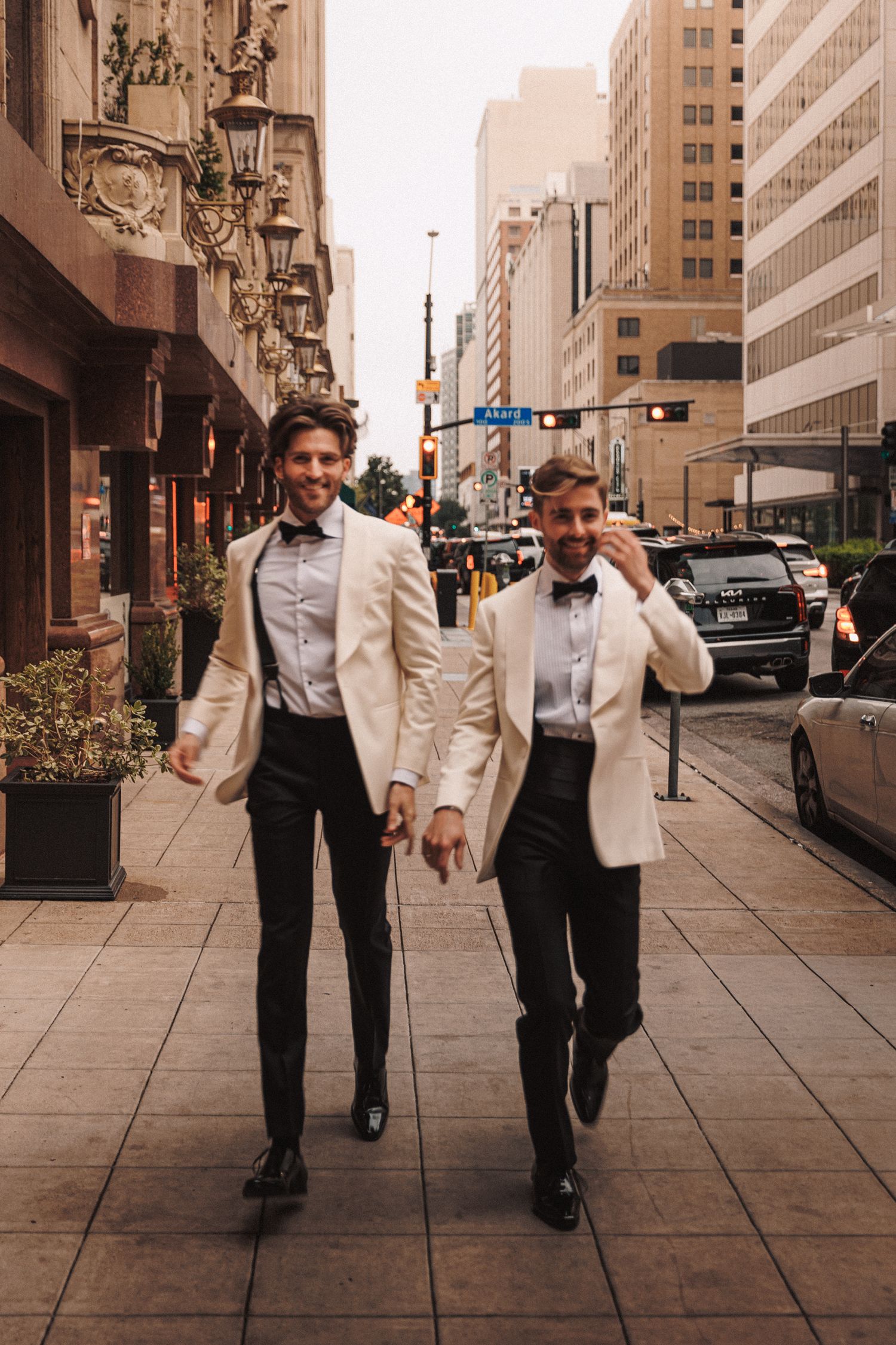 Two grooms in matching white blazers and black trousers walk hand-in-hand down a city street.