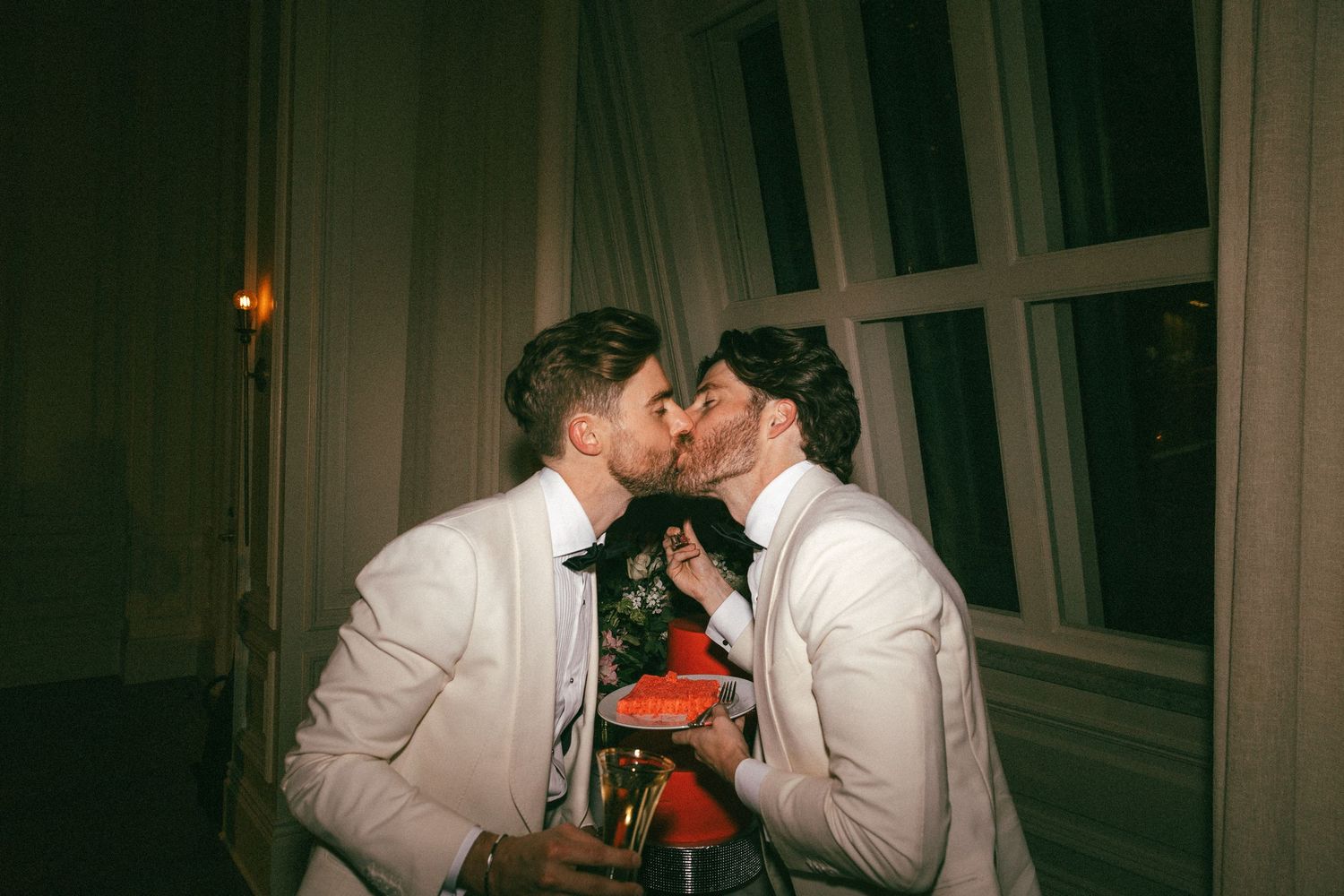 Two grooms in white tuxedos share a romantic kiss while holding red flowers at their wedding reception.