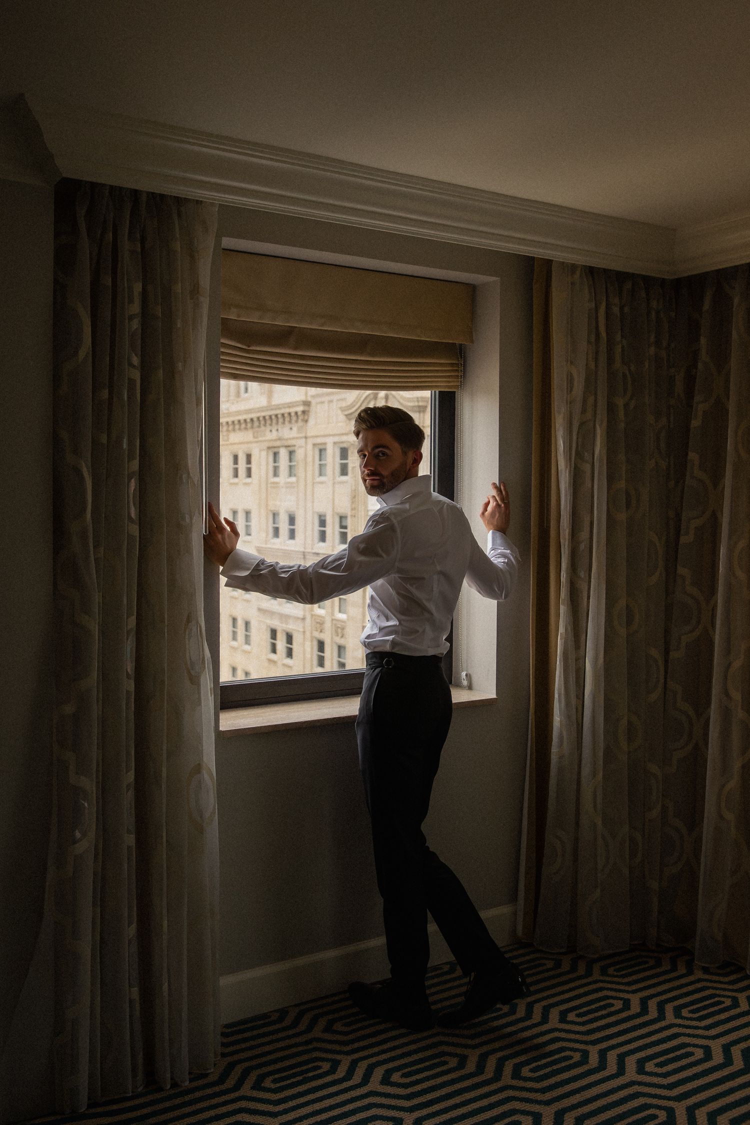Groom in white shirt standing between curtains at hotel window with urban skyline backdrop.