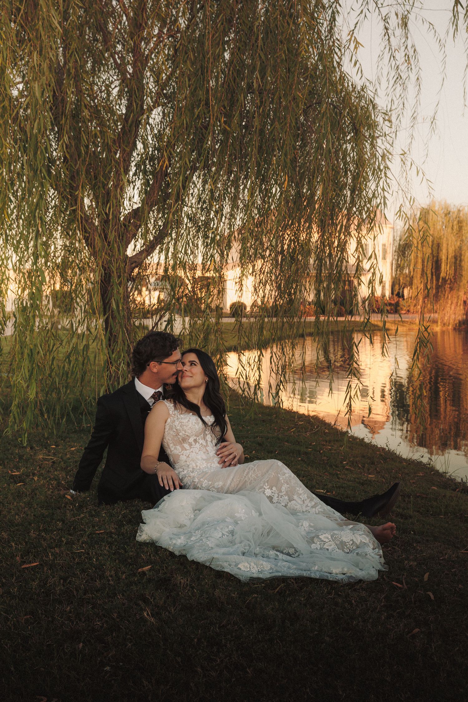 Bride and groom sitting together under weeping willow tree by reflective lake at sunset.