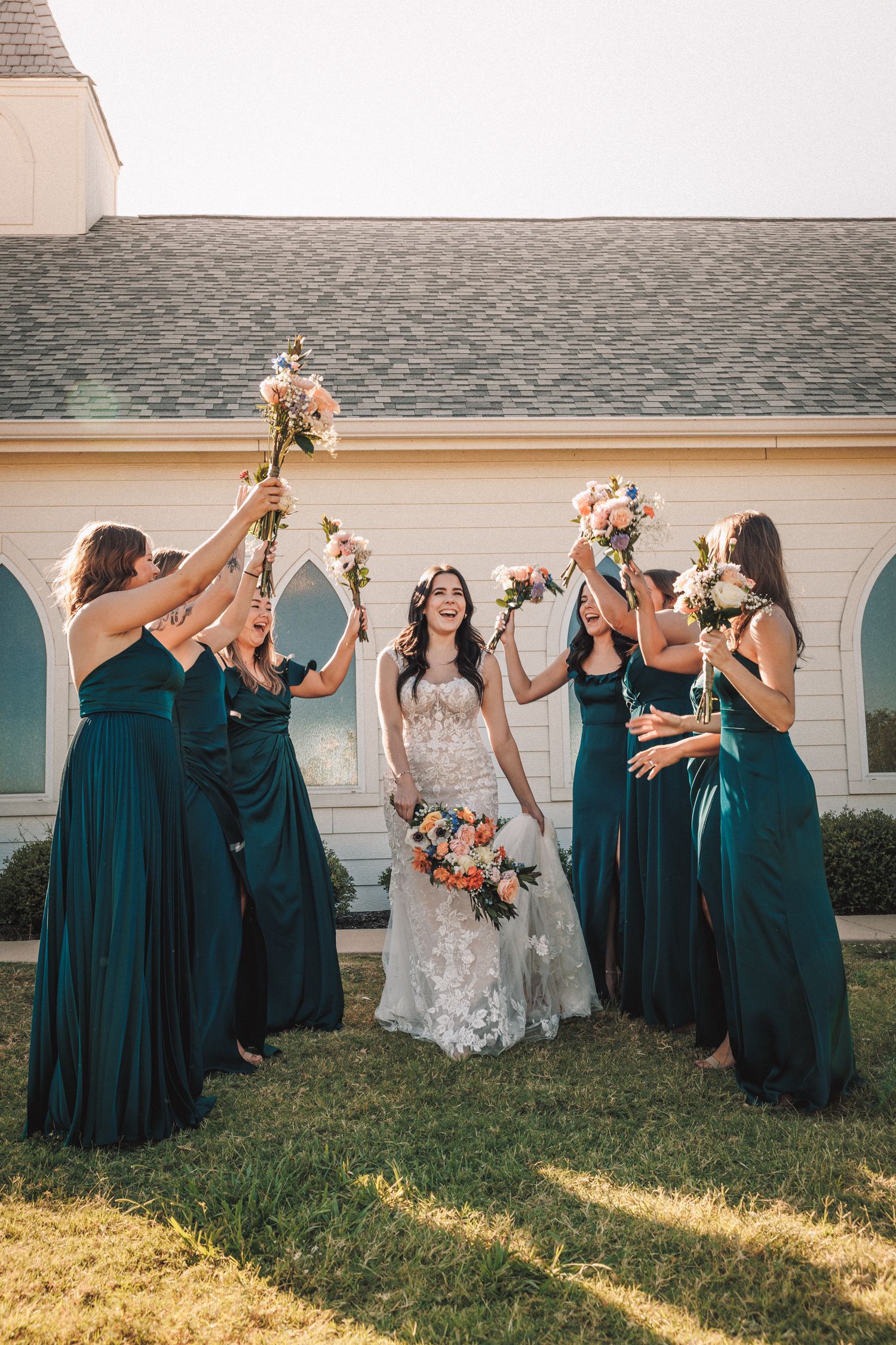 Bride in lace gown with bridesmaids in teal dresses raising bouquets outside a white chapel.