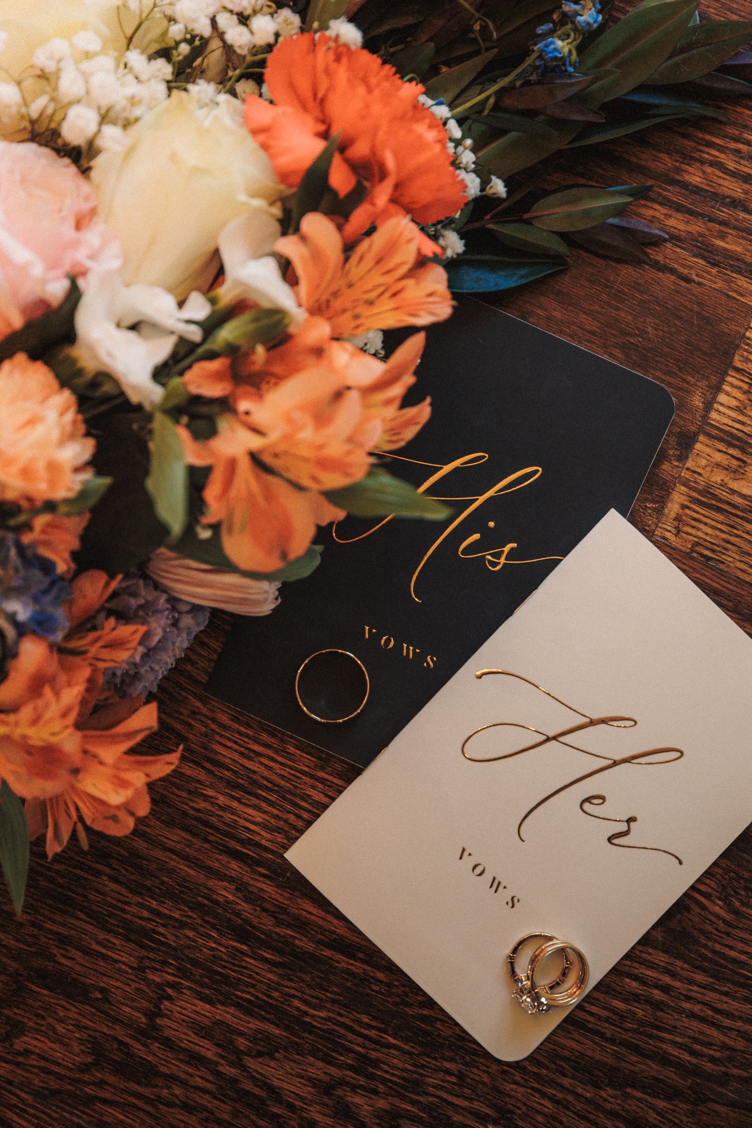His and her vows cards with wedding rings beside a colorful orange and pink bridal bouquet on a wooden surface.