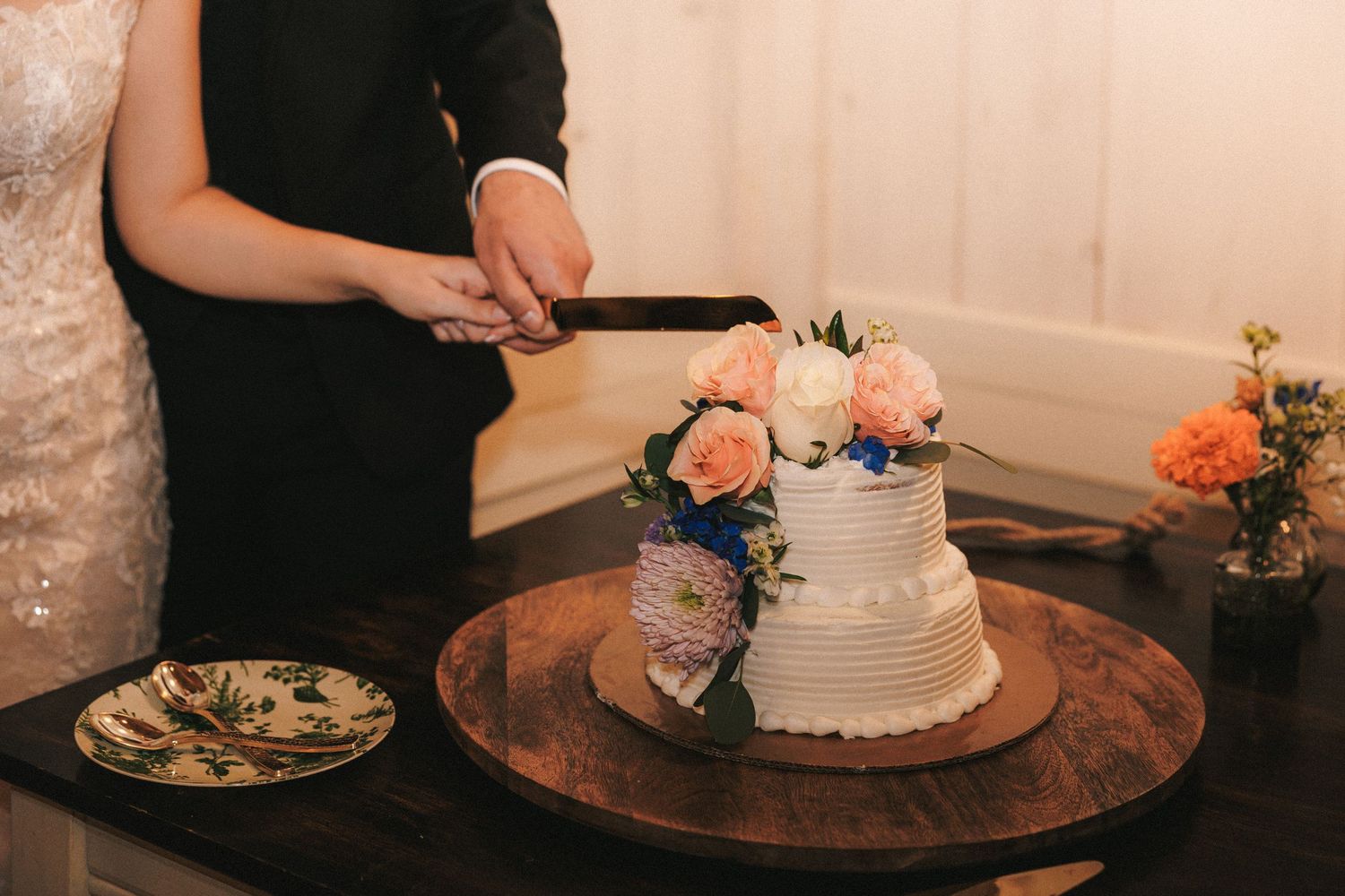 Bride and groom cutting elegant two-tier white wedding cake adorned with fresh pink roses.