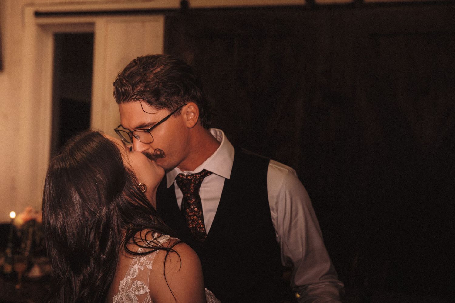 Groom with glasses and mustache shares a romantic kiss with bride in warm candlelit barn setting.