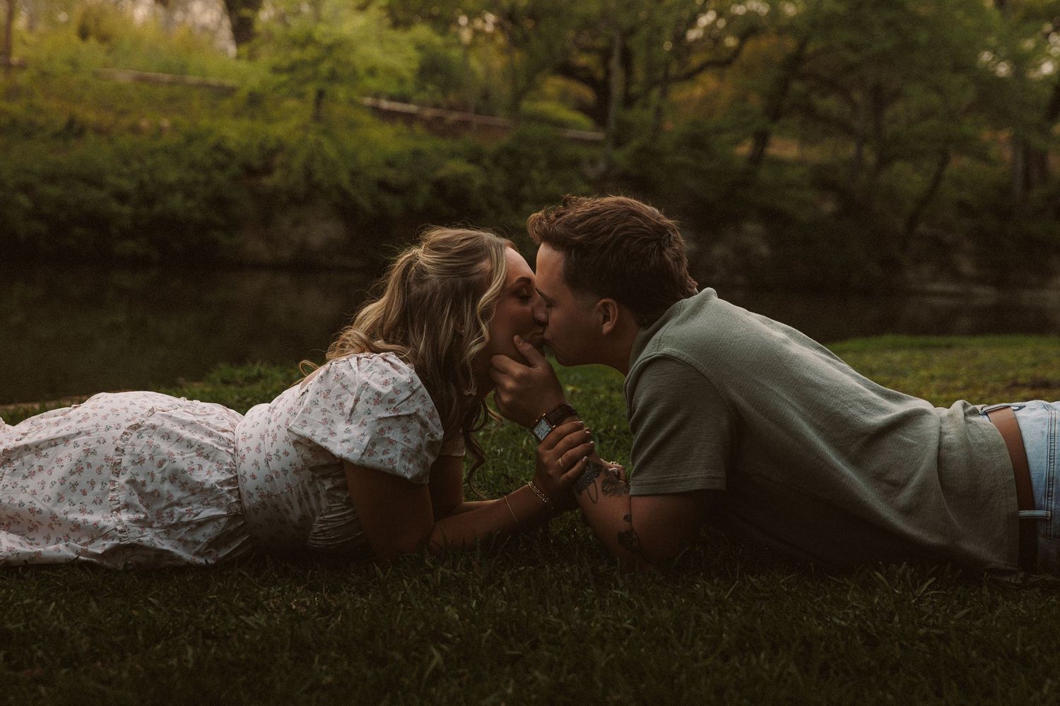 Couple lying on grass holding hands and sharing a kiss near a pond in a lush green park.