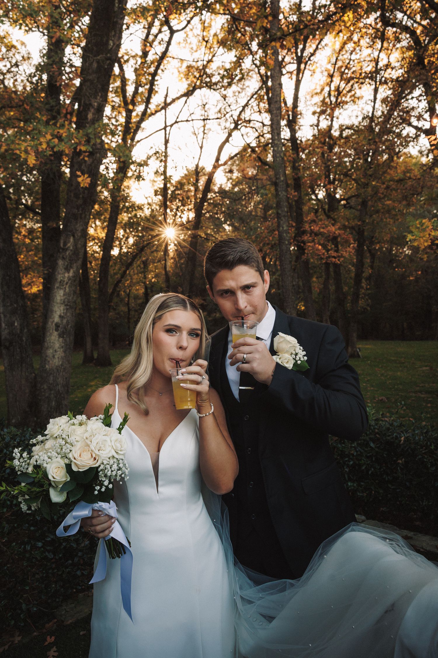 Bride and groom sipping drinks together in a sunlit autumn forest during their wedding celebration.