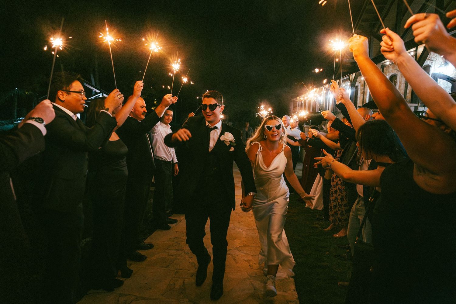 Newlyweds walk through sparkler exit tunnel surrounded by cheering wedding guests at night.