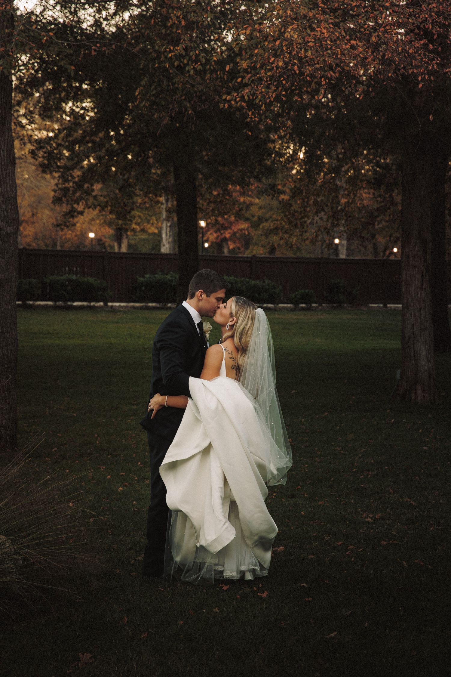 A bride and groom share a romantic kiss in a lush park at dusk during their wedding.