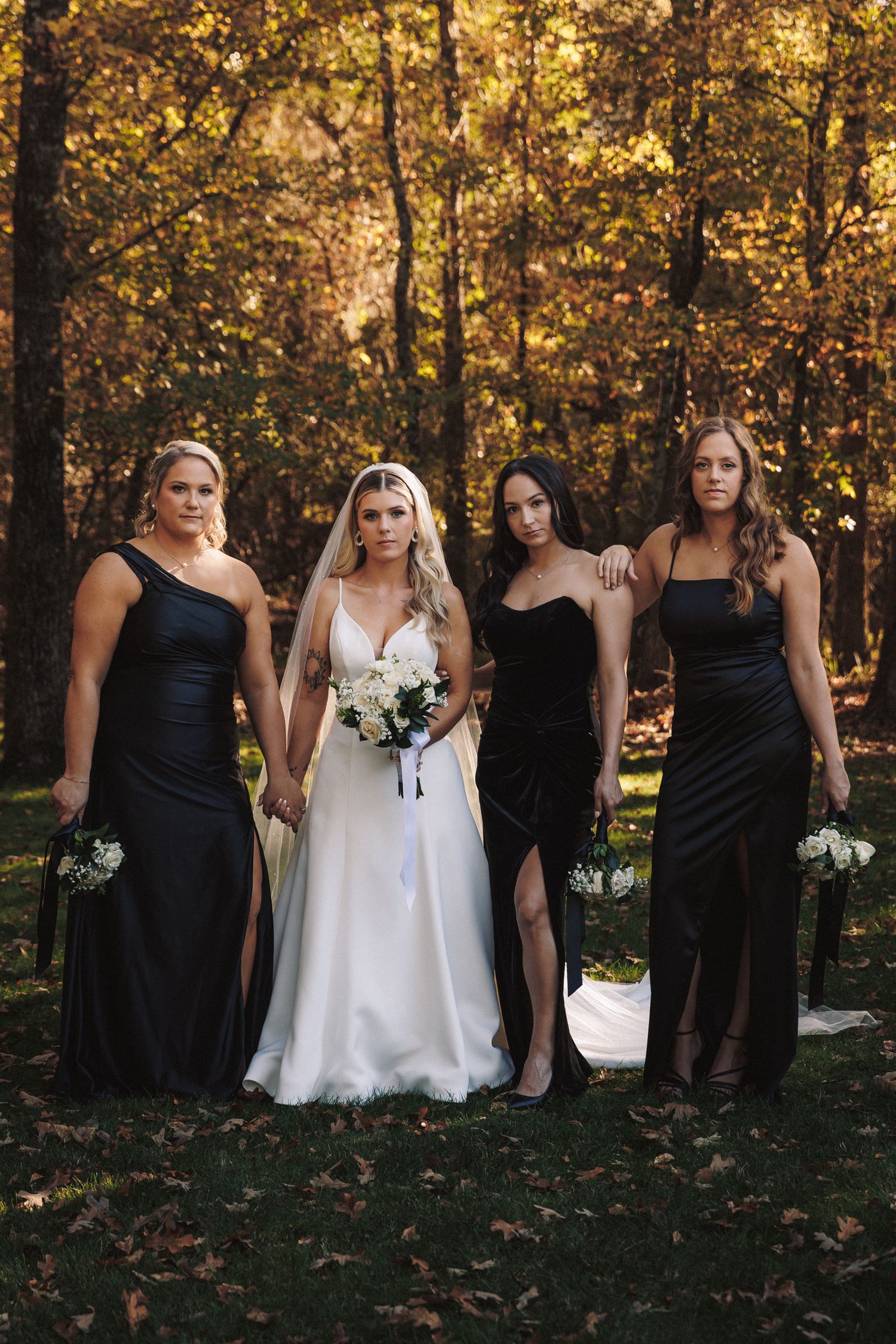 Bride in white gown with veil poses with three bridesmaids in black dresses amid autumn foliage.