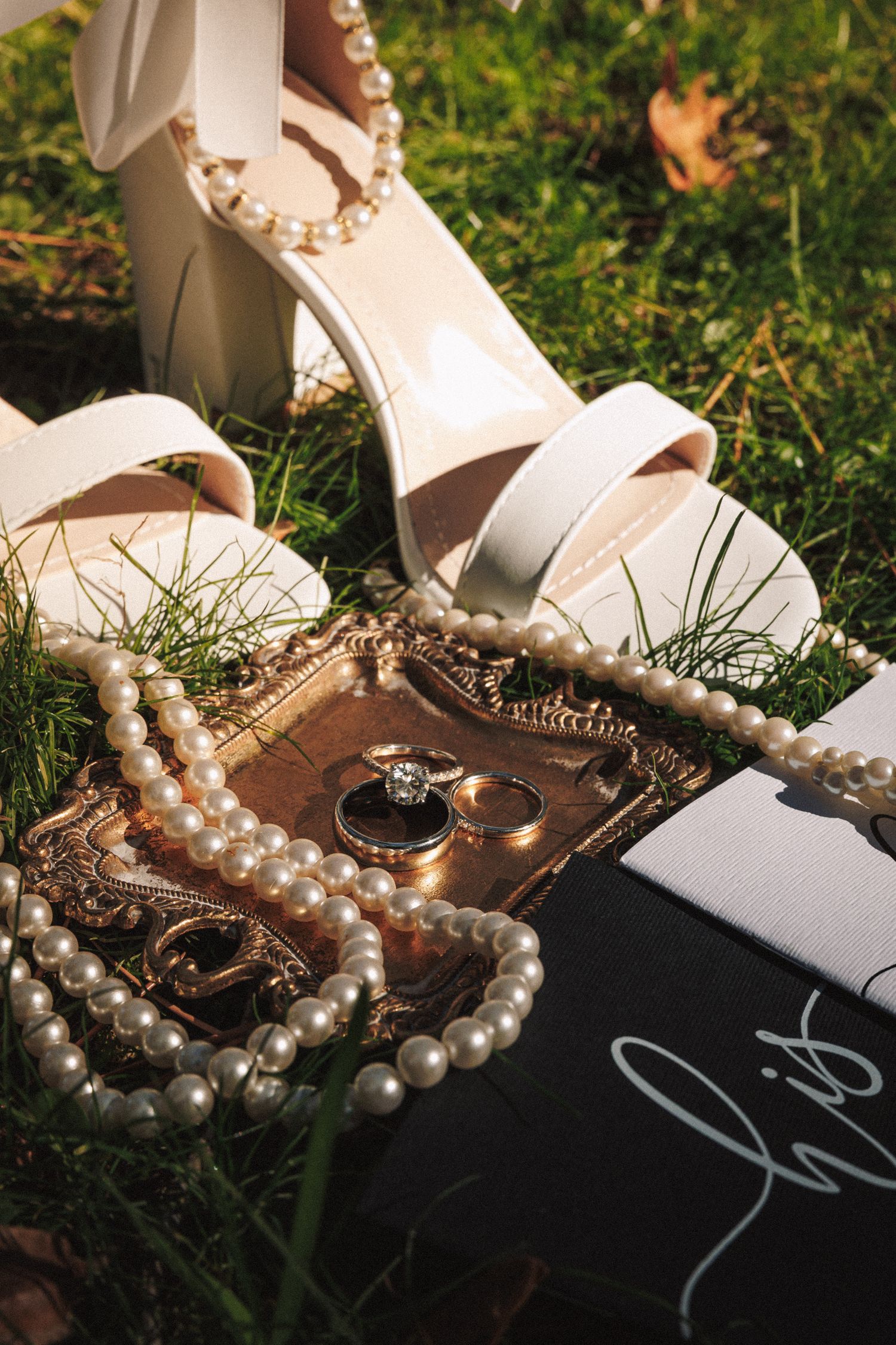 Bridal flat lay with white heels, pearl necklace, wedding rings on gold tray, and a his and hers card on green grass.