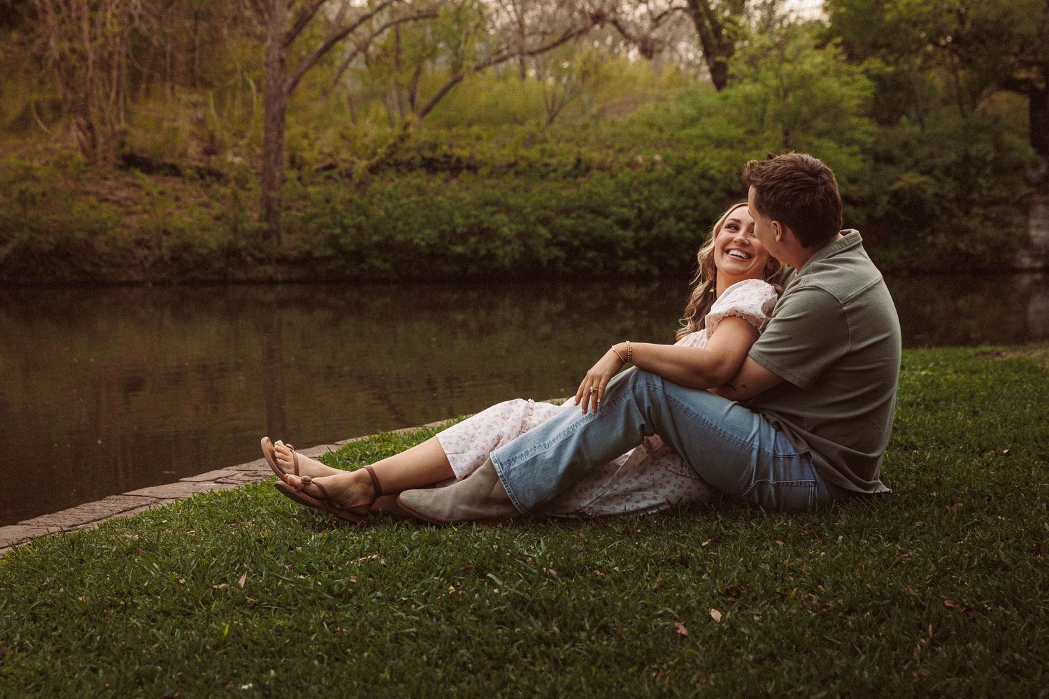 Happy couple sitting together on grass by a peaceful pond, laughing and embracing in a lush park.