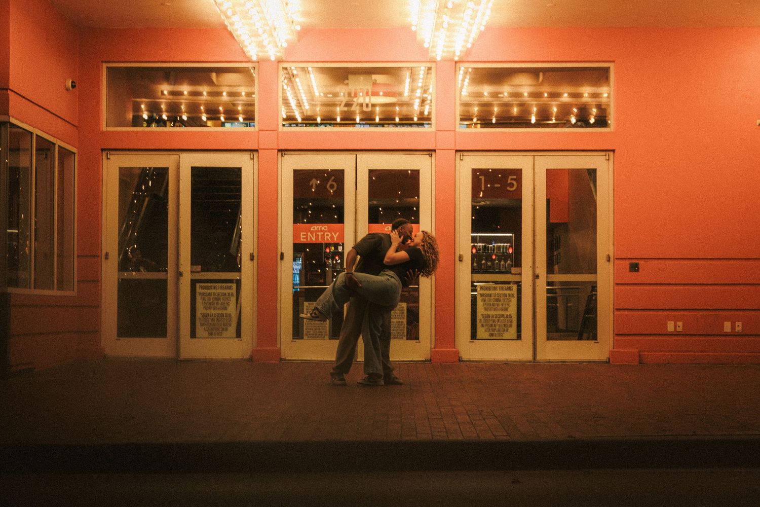 A couple embraces outside illuminated theater entrance doors at night with warm orange lighting.