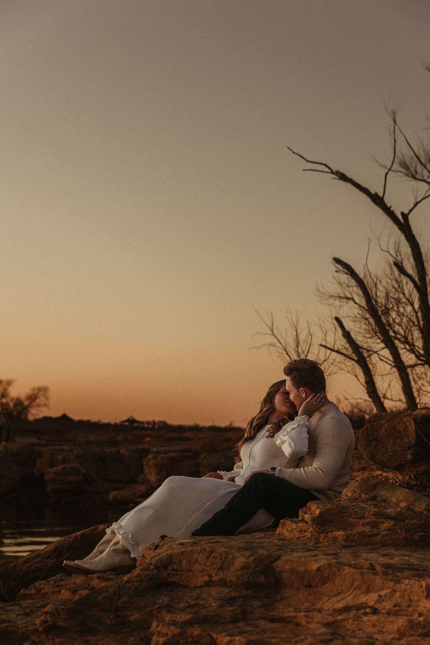 A couple resting together on rocky terrain at sunset with bare tree branches in the background.