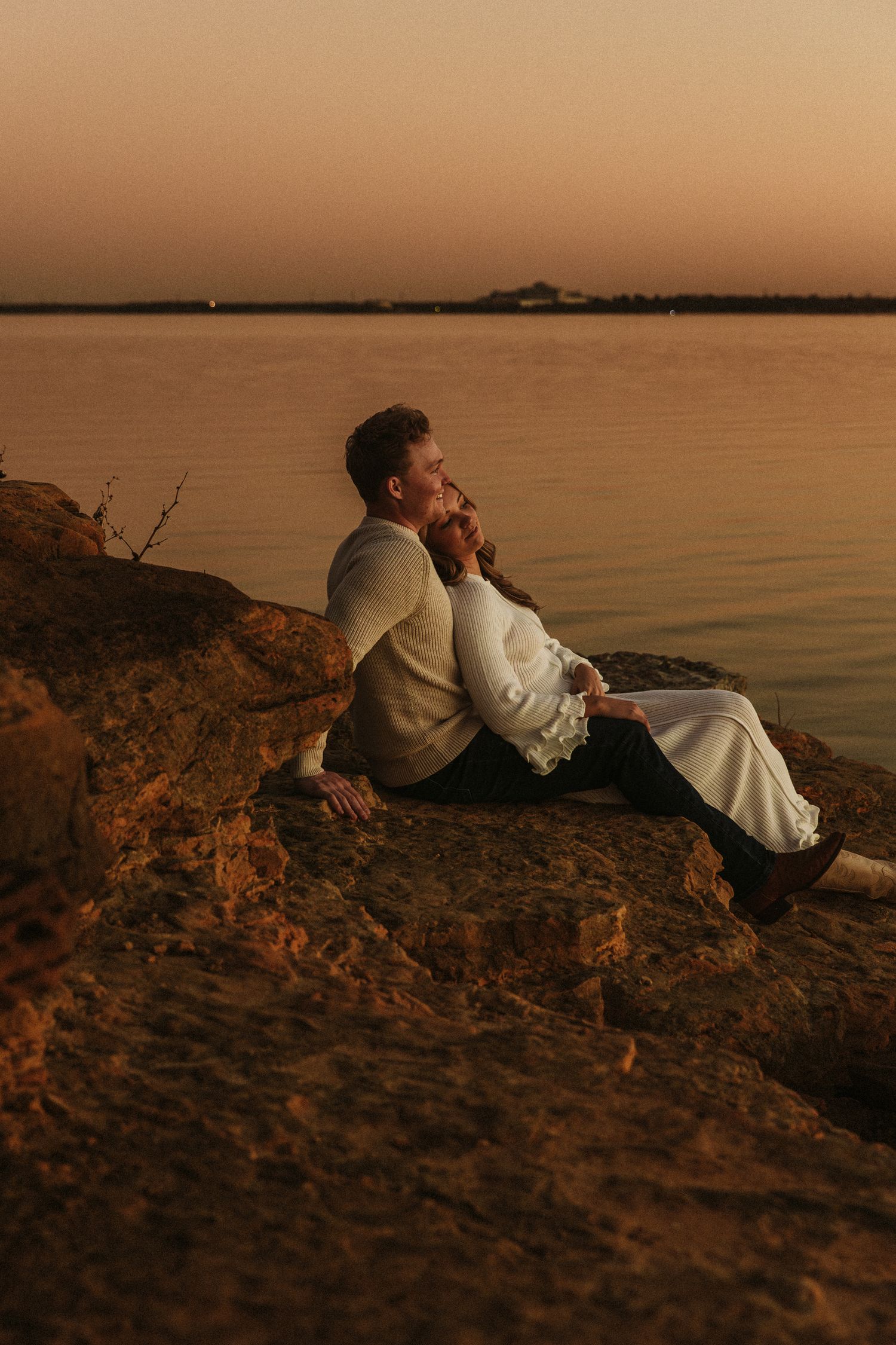 A couple sits together on a rocky cliff overlooking a serene lake at sunset.