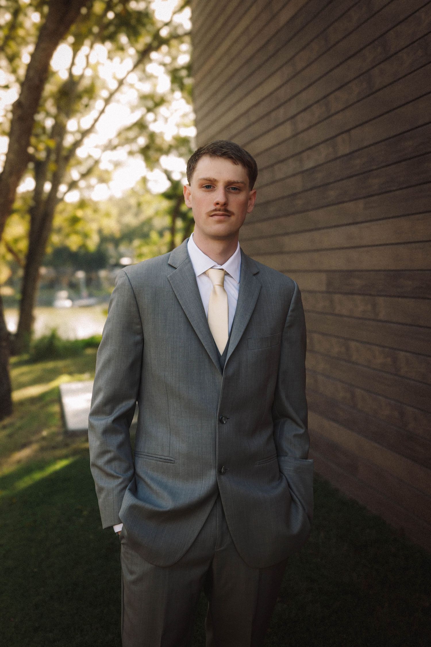 A portrait series of a person in a gray suit and white tie standing against a brown wooden wall outdoors.