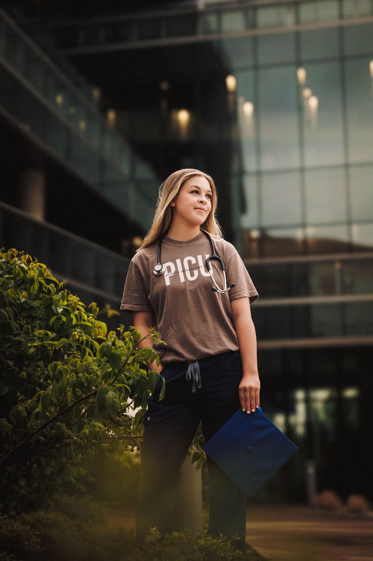 Recent graduate poses on university campus with modern architecture and evening lighting in background.