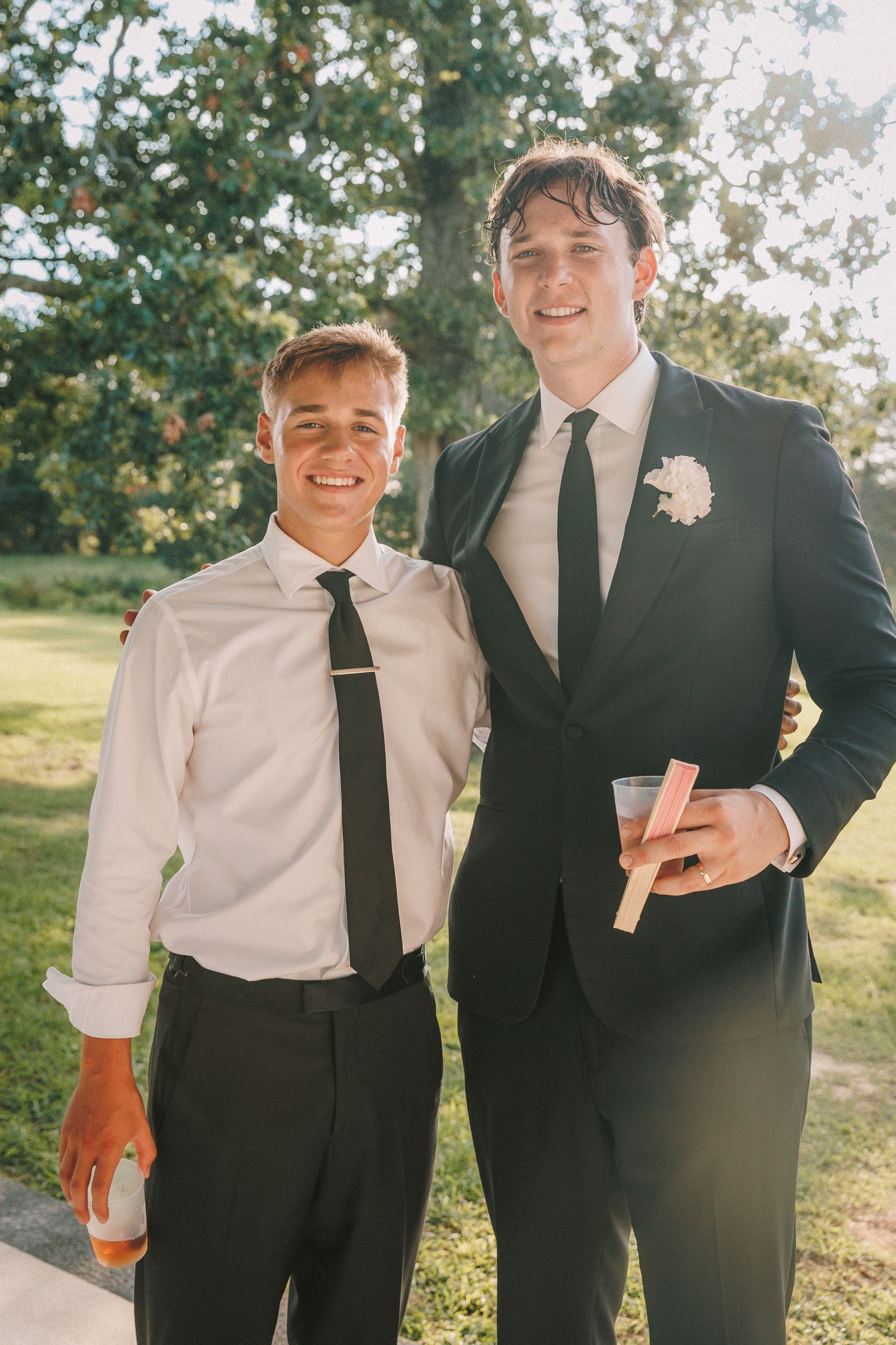 Two wedding attendees share a laugh while holding drinks at an outdoor reception.