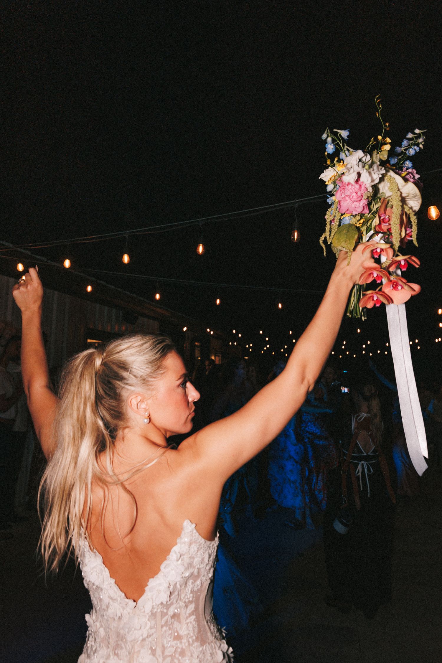 Wedding celebration with bride holding bouquet overhead under string lights.