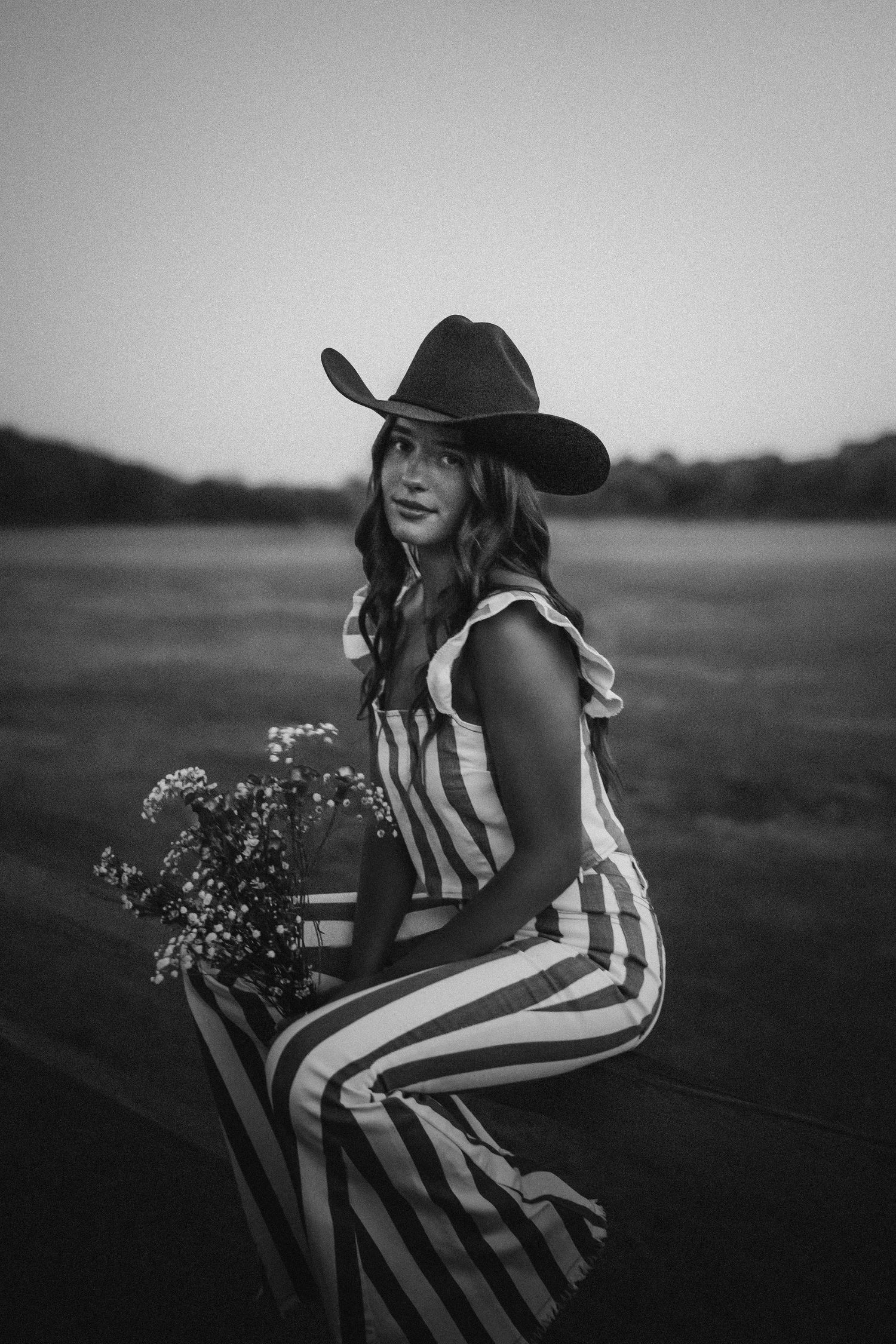 A figure in a black and white striped dress and wide-brimmed hat poses with wildflowers in a monochrome outdoor setting.