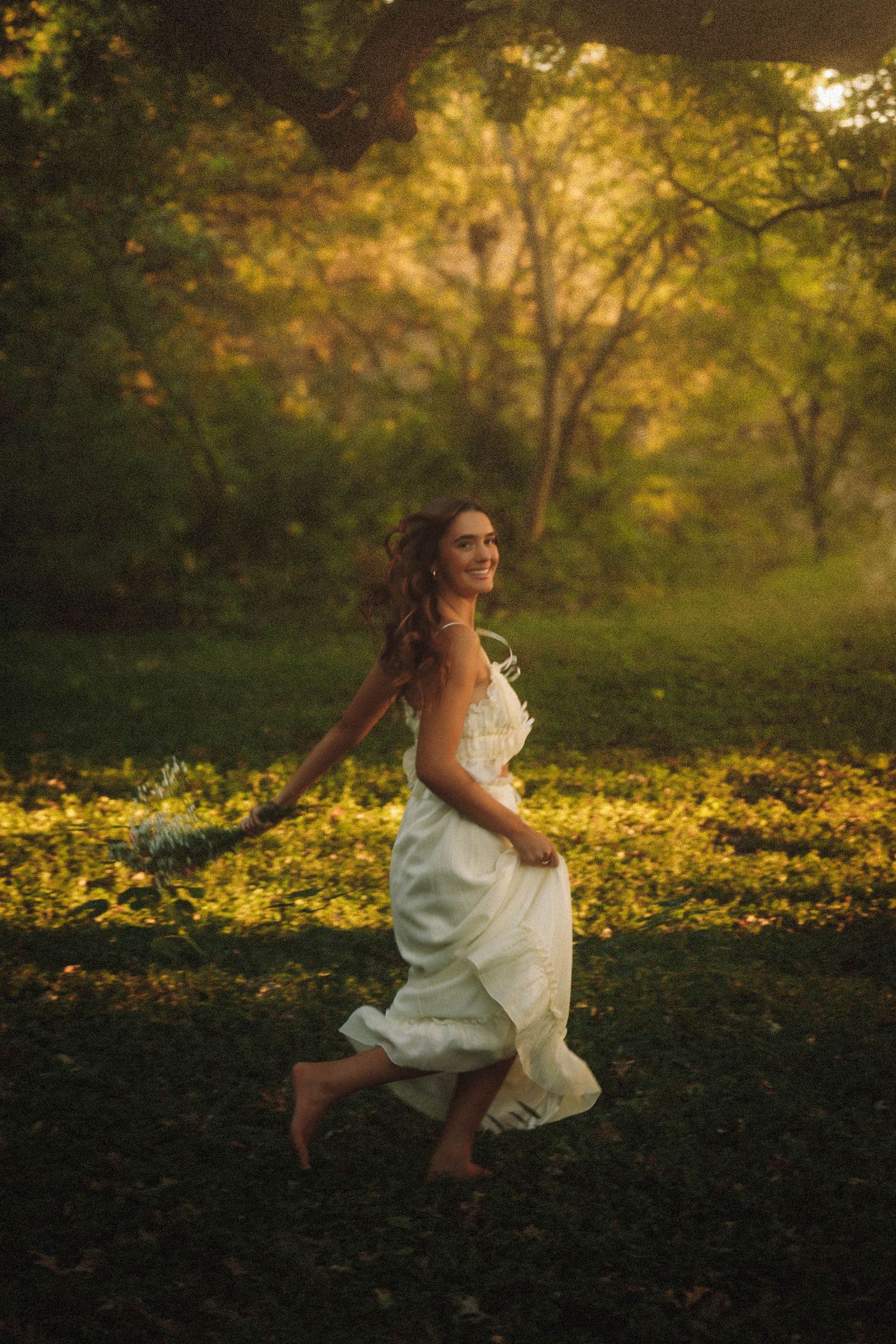 A figure in a flowing white dress poses gracefully in a sunlit forest meadow with yellow wildflowers in the background.