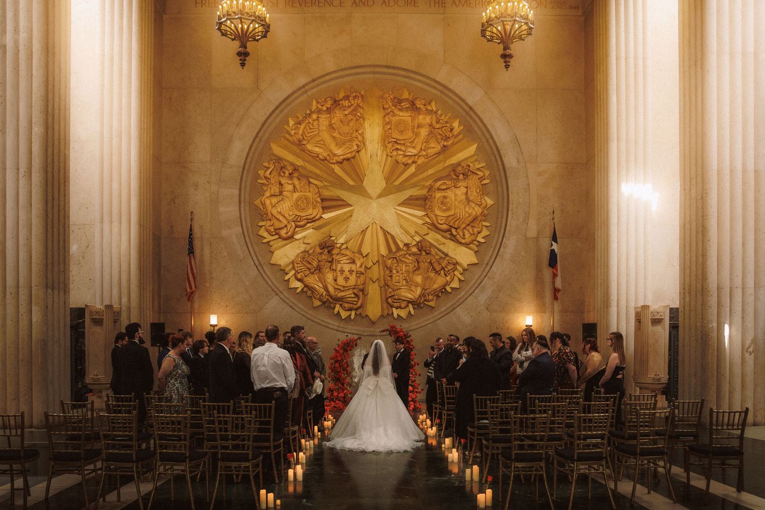 Wedding ceremony in grand hall with golden circular mural, candlelit aisle and seated guests.