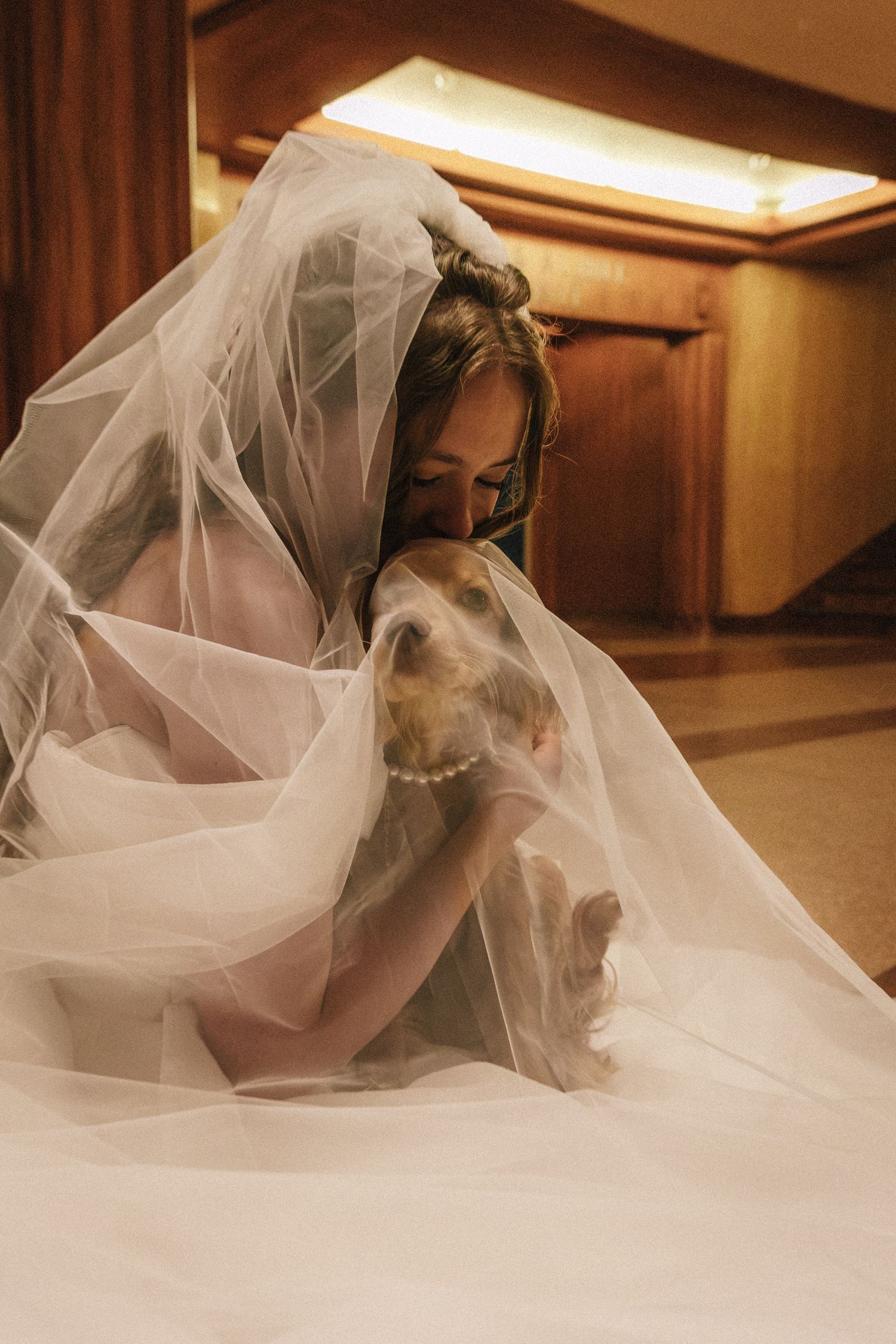 Bride in white gown and veil cuddling with a dog on the floor indoors.