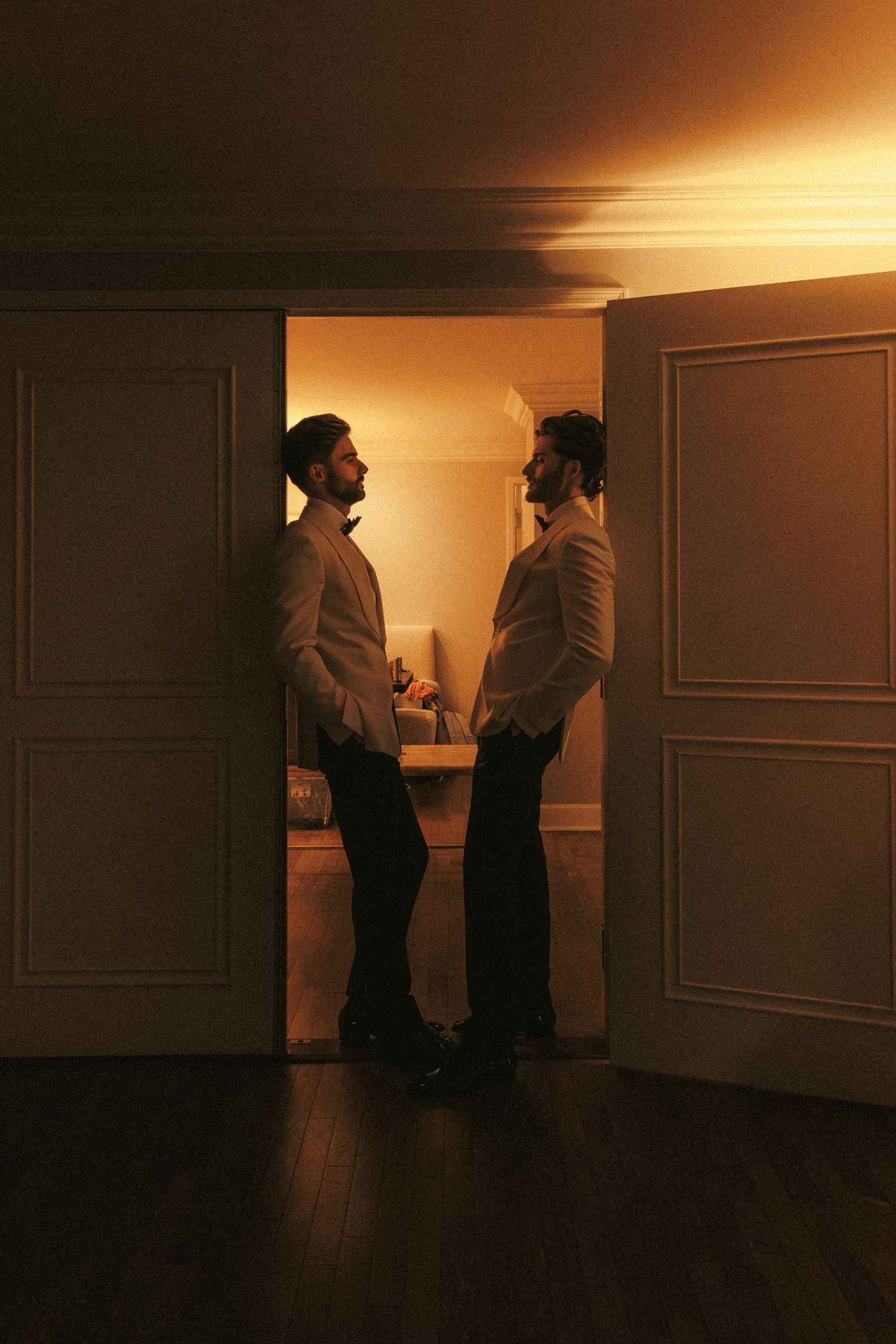 Two grooms in white tuxedos face each other in a warmly lit doorway on their wedding day.