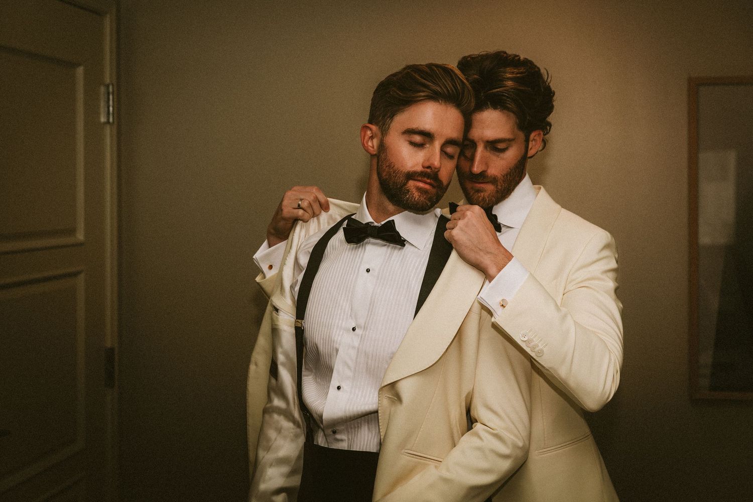 Two grooms in ivory tuxedos with bow ties share an intimate moment while adjusting each other's jackets.