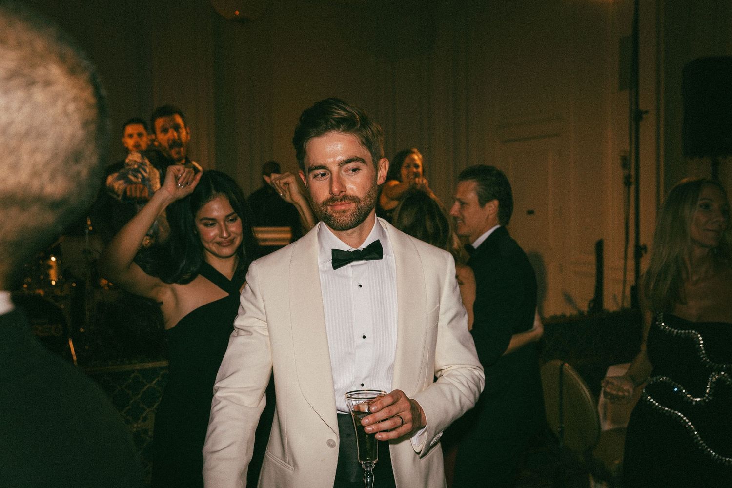Stylish groom in cream tuxedo holding a drink at an upscale wedding reception party.