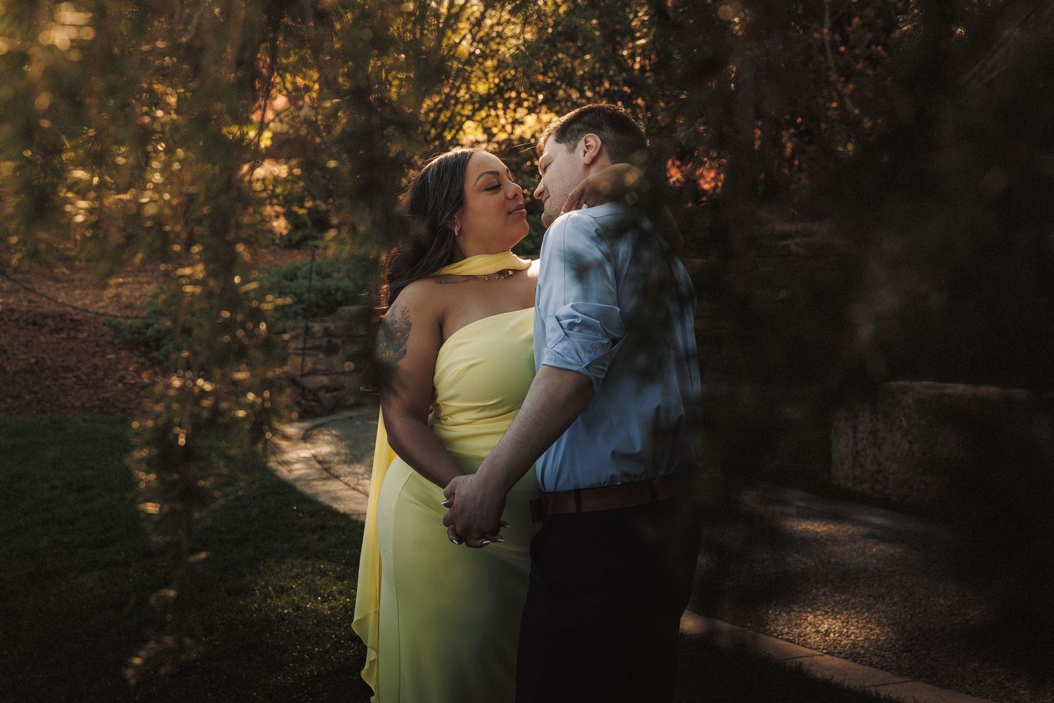 Couple sharing a romantic kiss in a sunlit garden, woman in yellow gown and man in blue shirt.