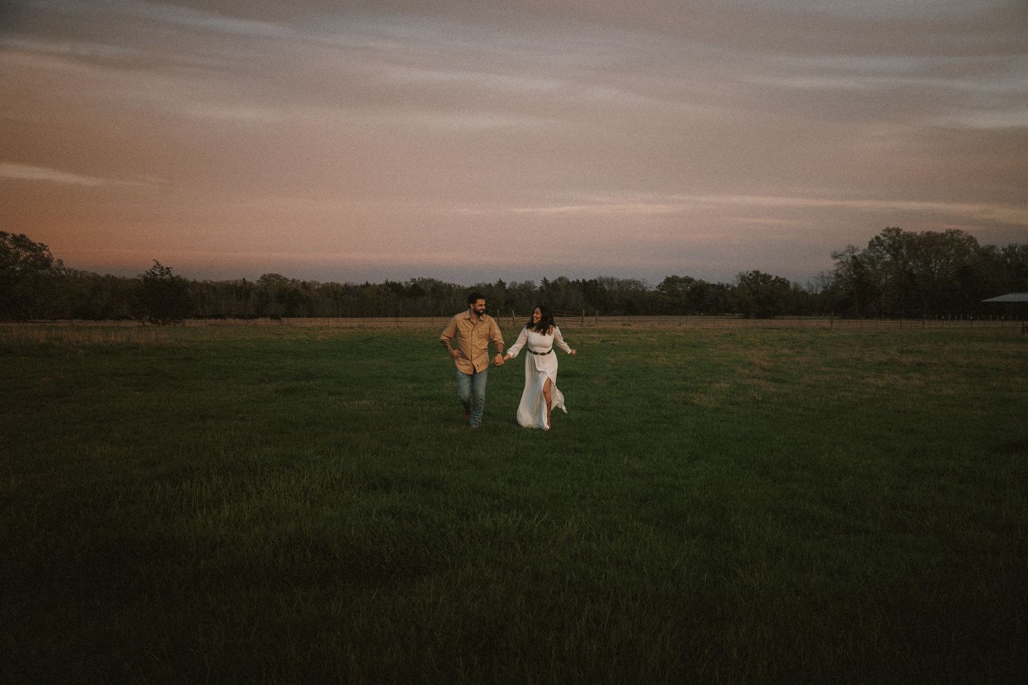 Couple holding hands walking through a lush green field at sunset with a colorful sky and trees in the background.