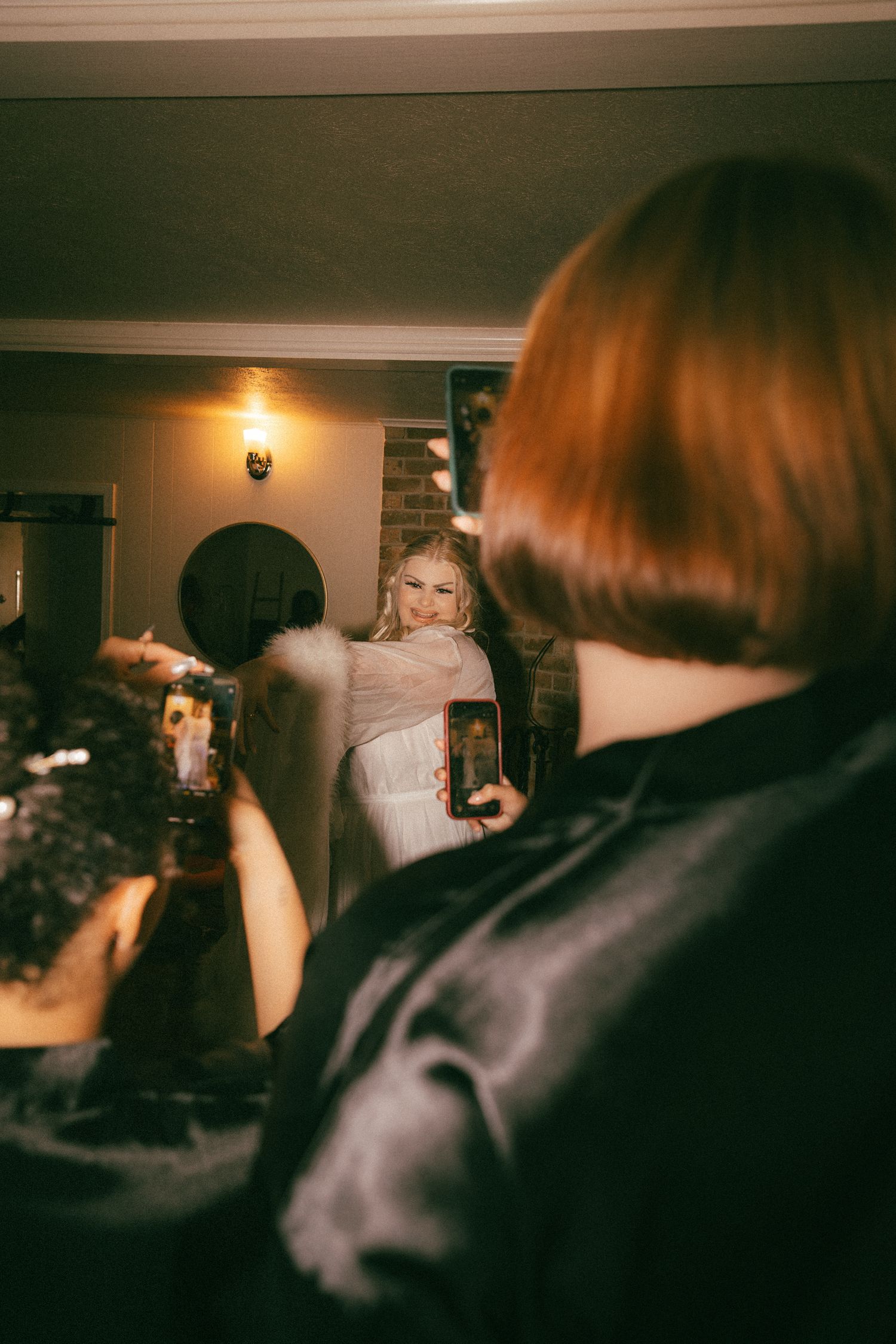 Wedding guests photographing a smiling blonde bride in a white gown with feather wrap at an indoor venue.