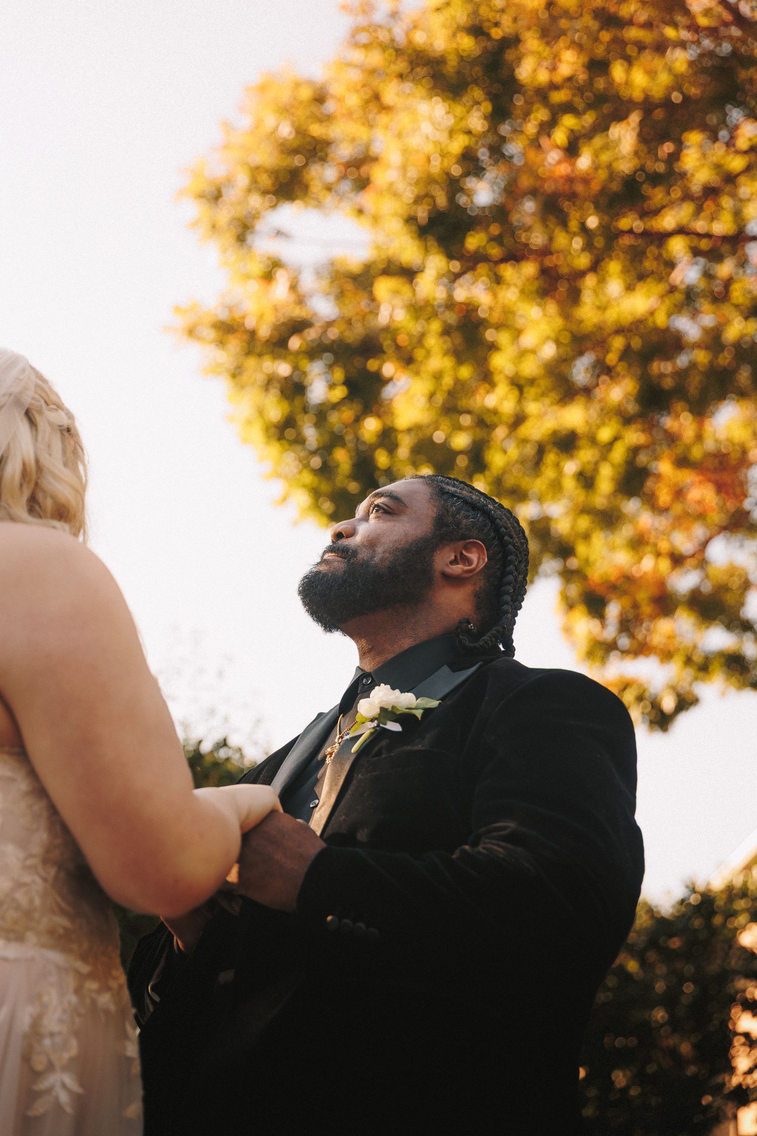 Groom in black suit with braids gazing upward during outdoor fall wedding ceremony.
