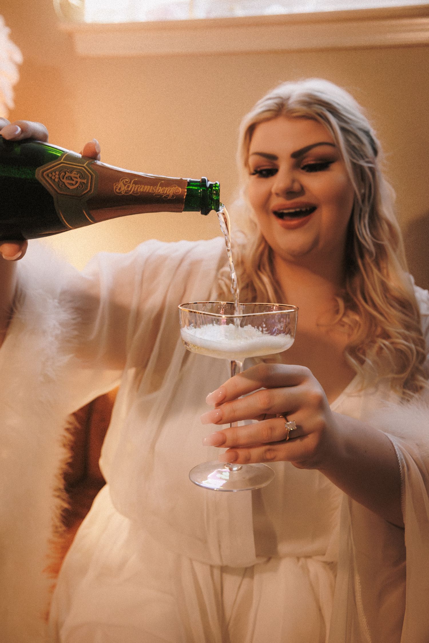 Excited bride in white robe holding coupe glass while champagne is poured during wedding morning preparations.