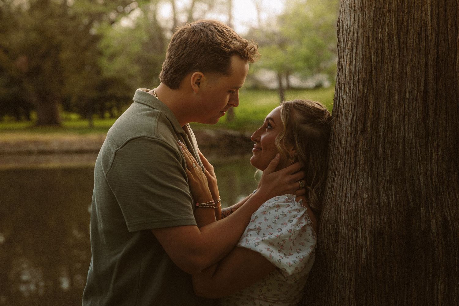 A romantic couple shares an intimate moment leaning against a tree by a sunlit lake.
