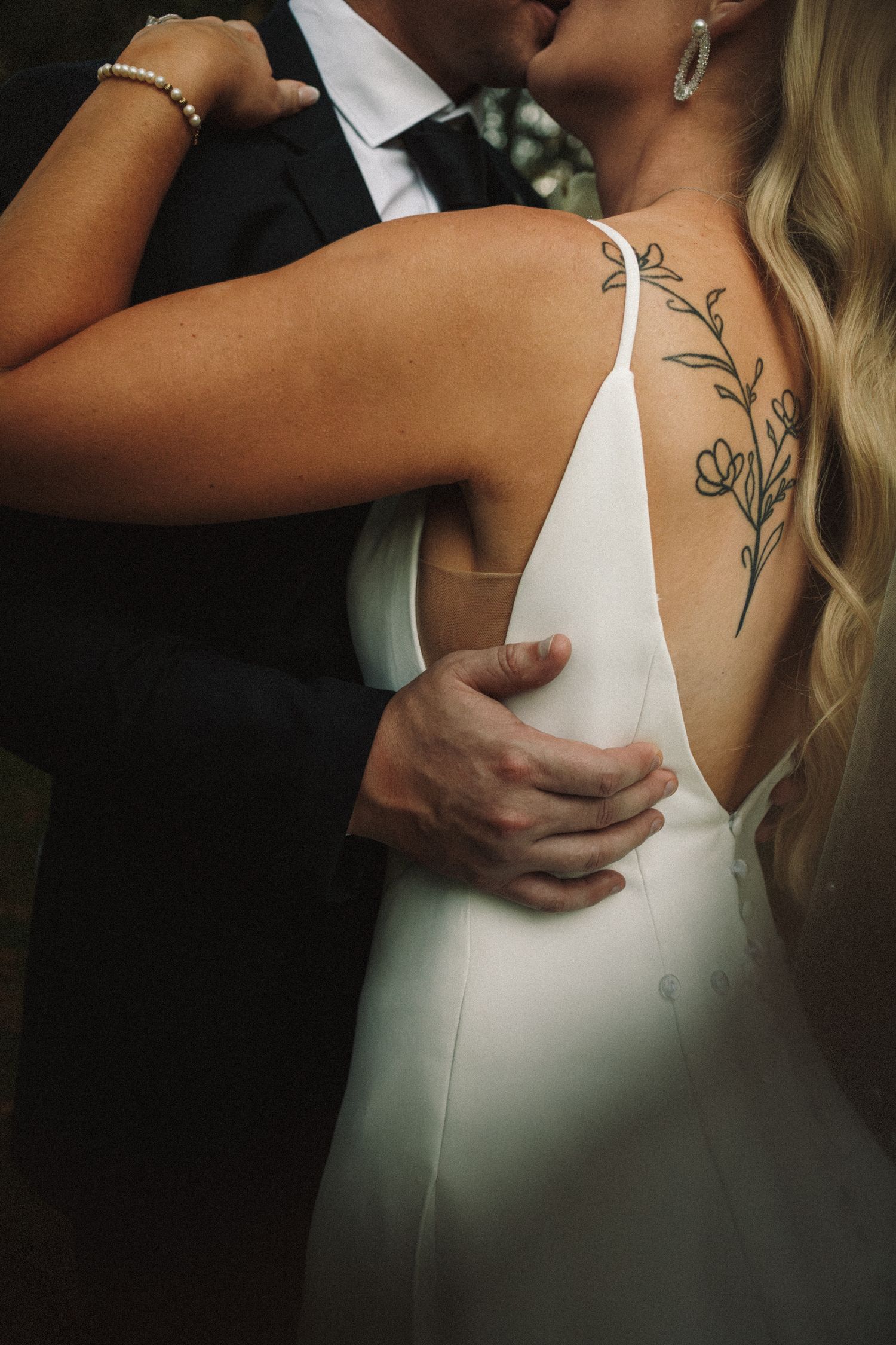 Close-up of bride with floral back tattoo embracing groom in black suit during romantic moment.