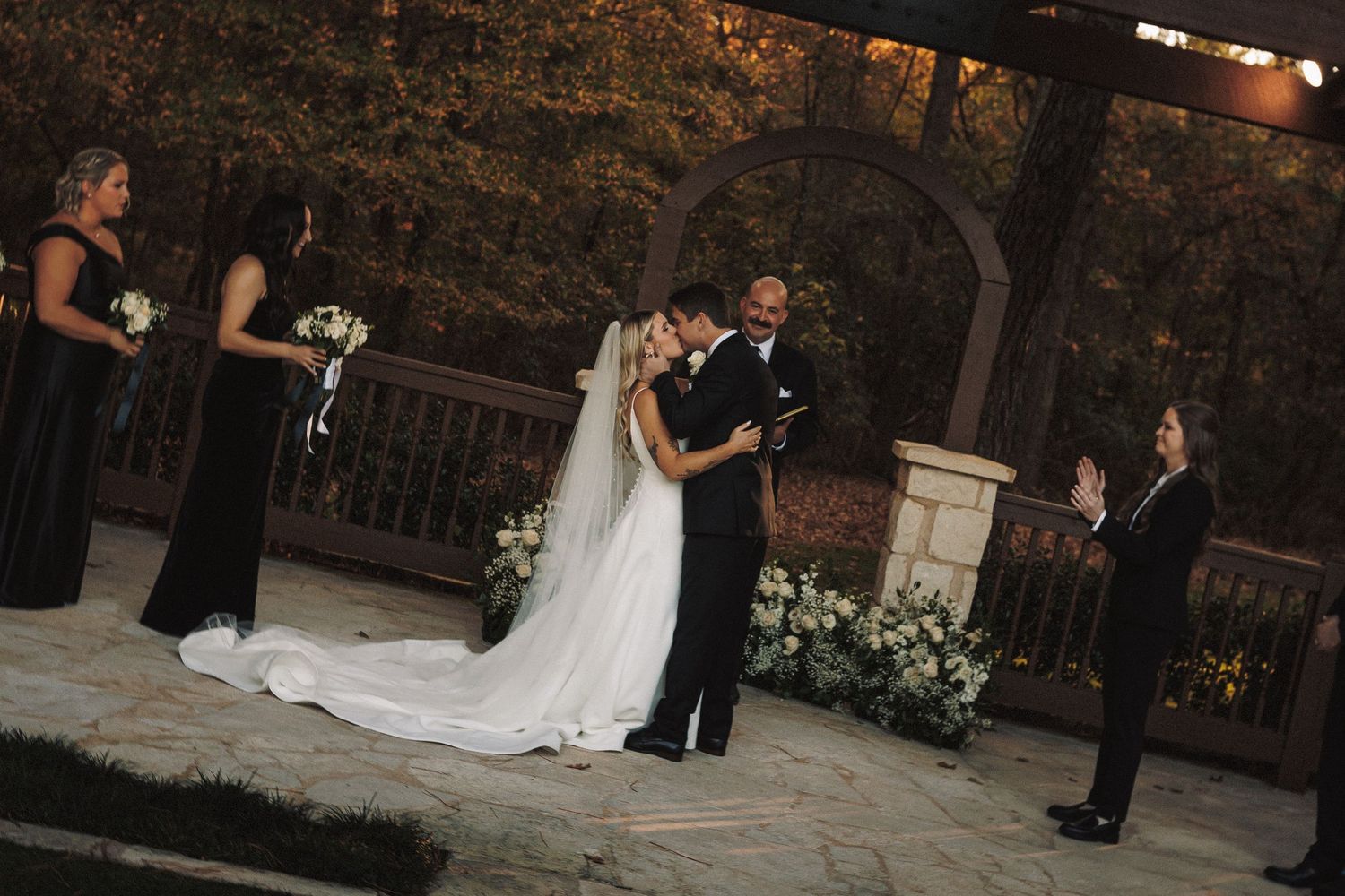 Bride and groom share first kiss at outdoor evening wedding ceremony as bridesmaids in black dresses look on.