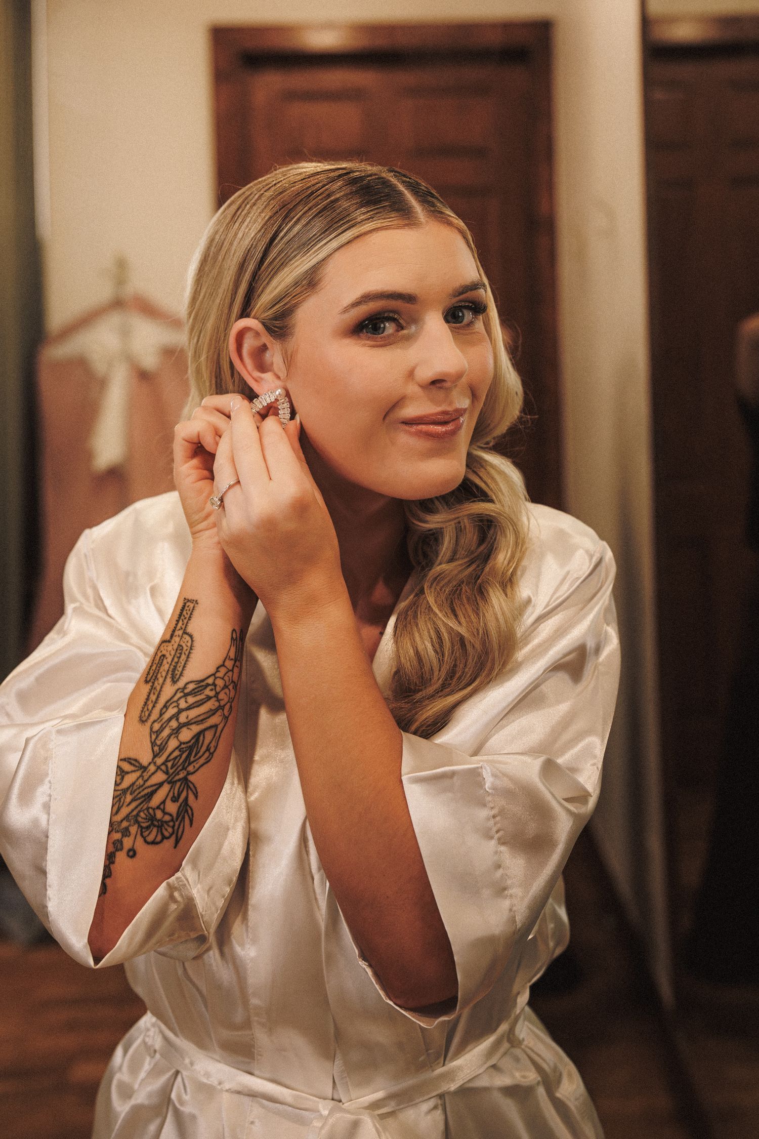 Beautiful blonde bride in white satin robe putting on earrings with tattoos visible on her arm.
