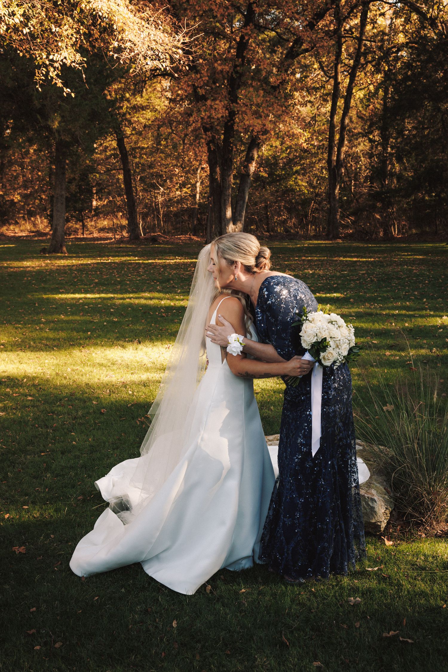 Bride in white gown and veil shares tender embrace with bridesmaid in navy sequin dress outdoors.