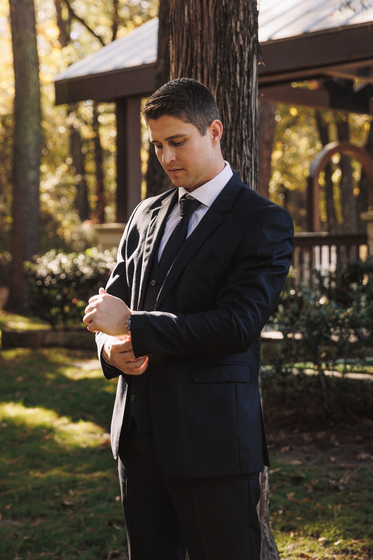 Groom in black suit adjusting cufflinks outdoors near a tree in warm autumn sunlight.