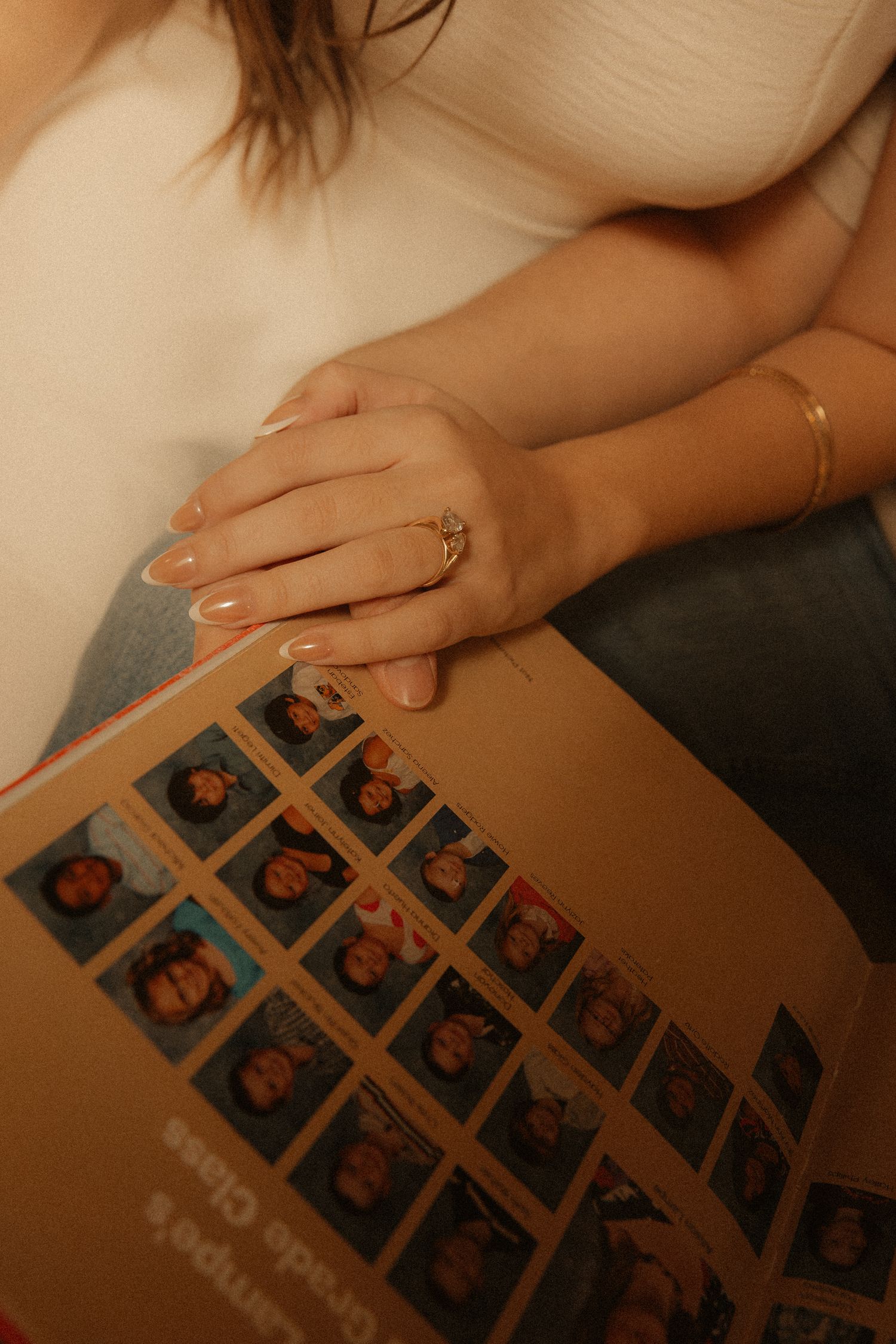 Woman holding a yearbook with photo strips, wearing a gold ring and bracelet, dressed in white top and jeans.