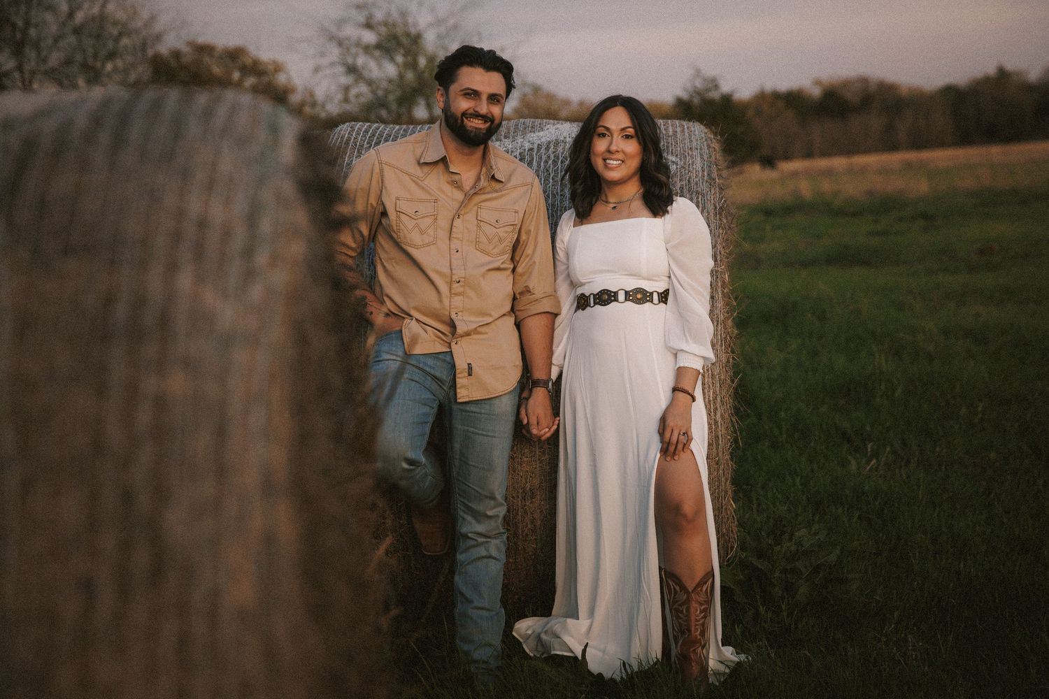 Couple posing by hay bales at sunset, woman in white dress, man in tan shirt.