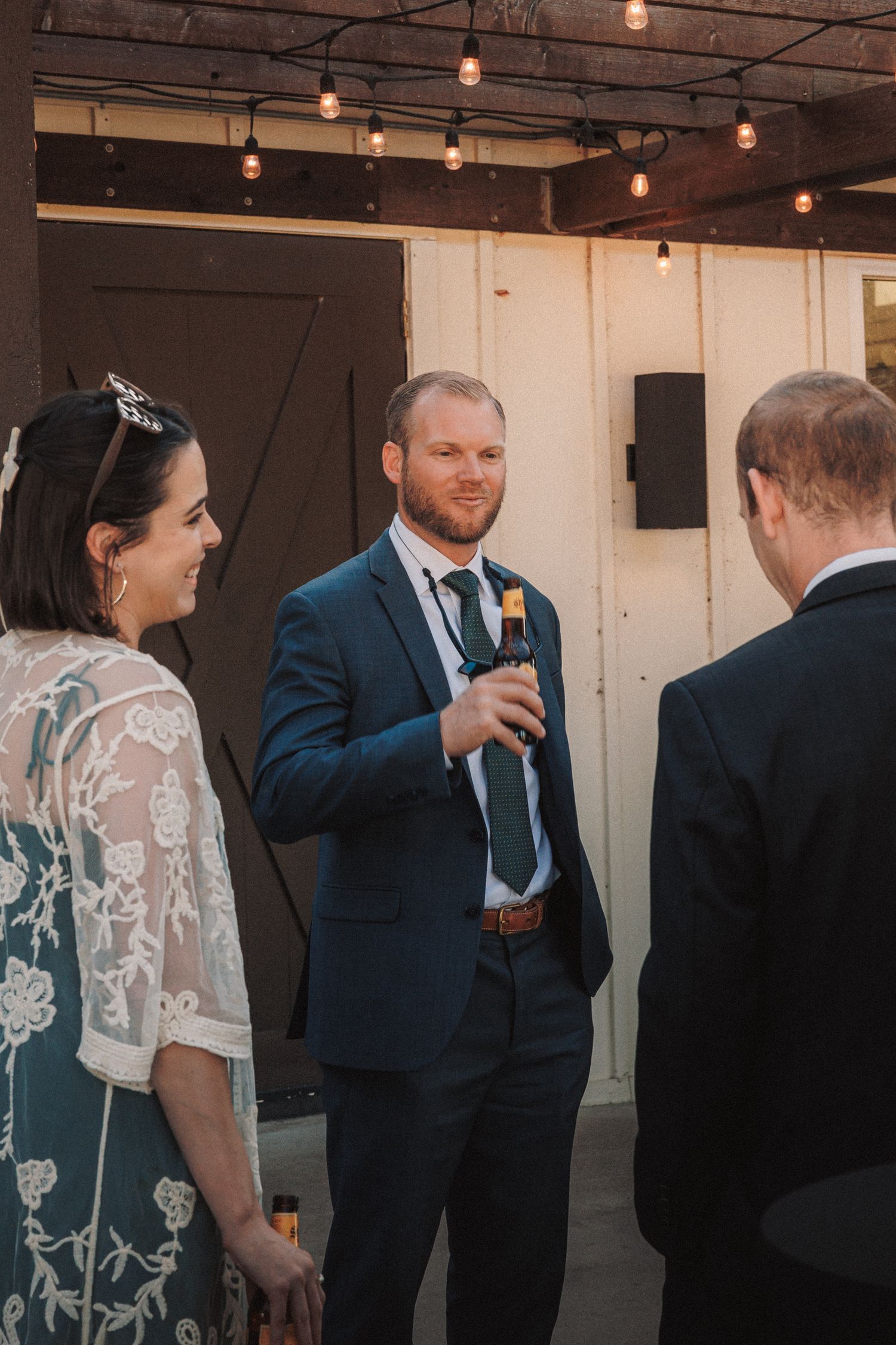 Bearded groom in navy suit holds a beer bottle while chatting with guests at an outdoor wedding reception.